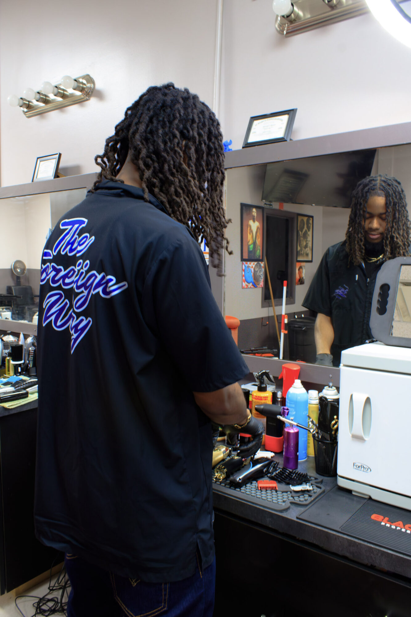 Person with curly hair working at a cluttered salon station, mirror reflecting their face, various hair products and tools visible