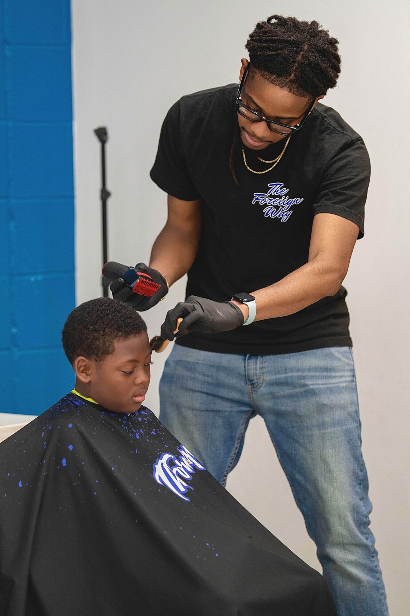 Barber cutting child's hair in a salon, child wearing a cape, barber wearing glasses and gloves, plain background.