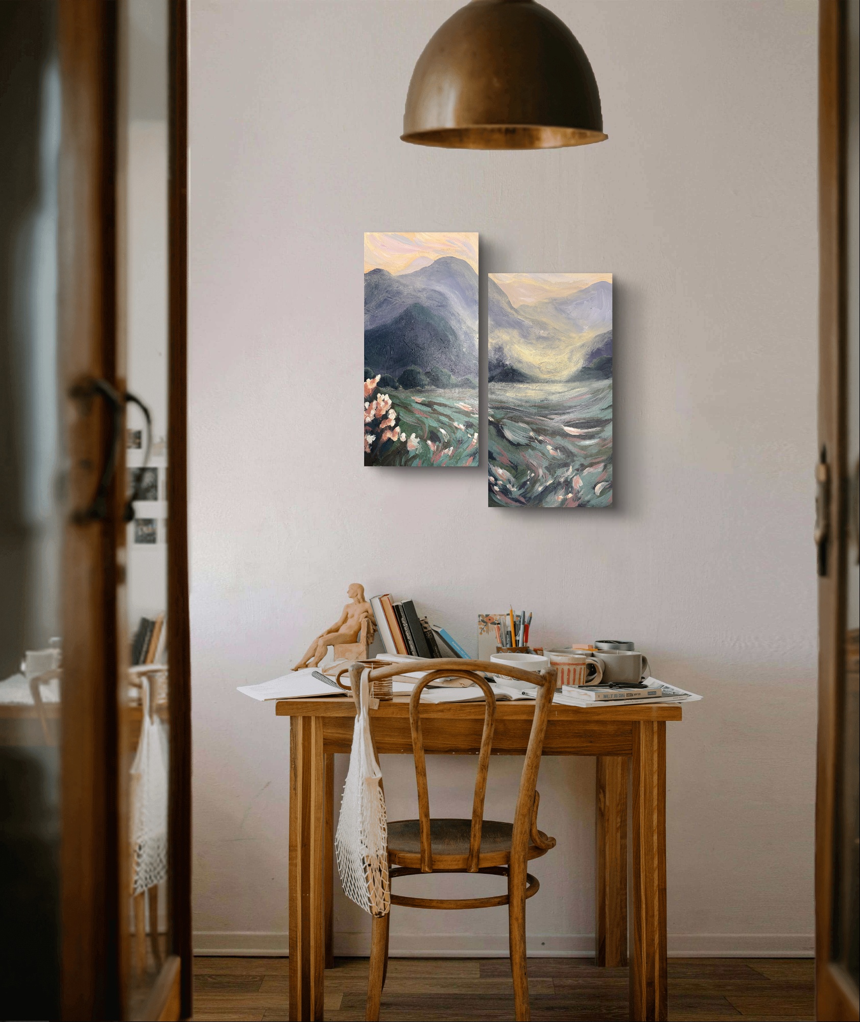 View of a small wooden desk with a chair, books, and a cup, against a white wall with two landscape paintings, a hanging lamp above, and a lace cloth on the desk.