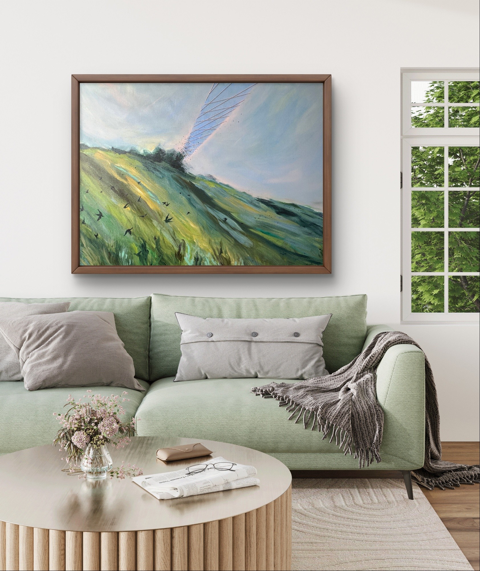Living room with a green sofa, pillows, a blanket, a round coffee table with books and flowers, a large framed painting, and a window with greenery outside.