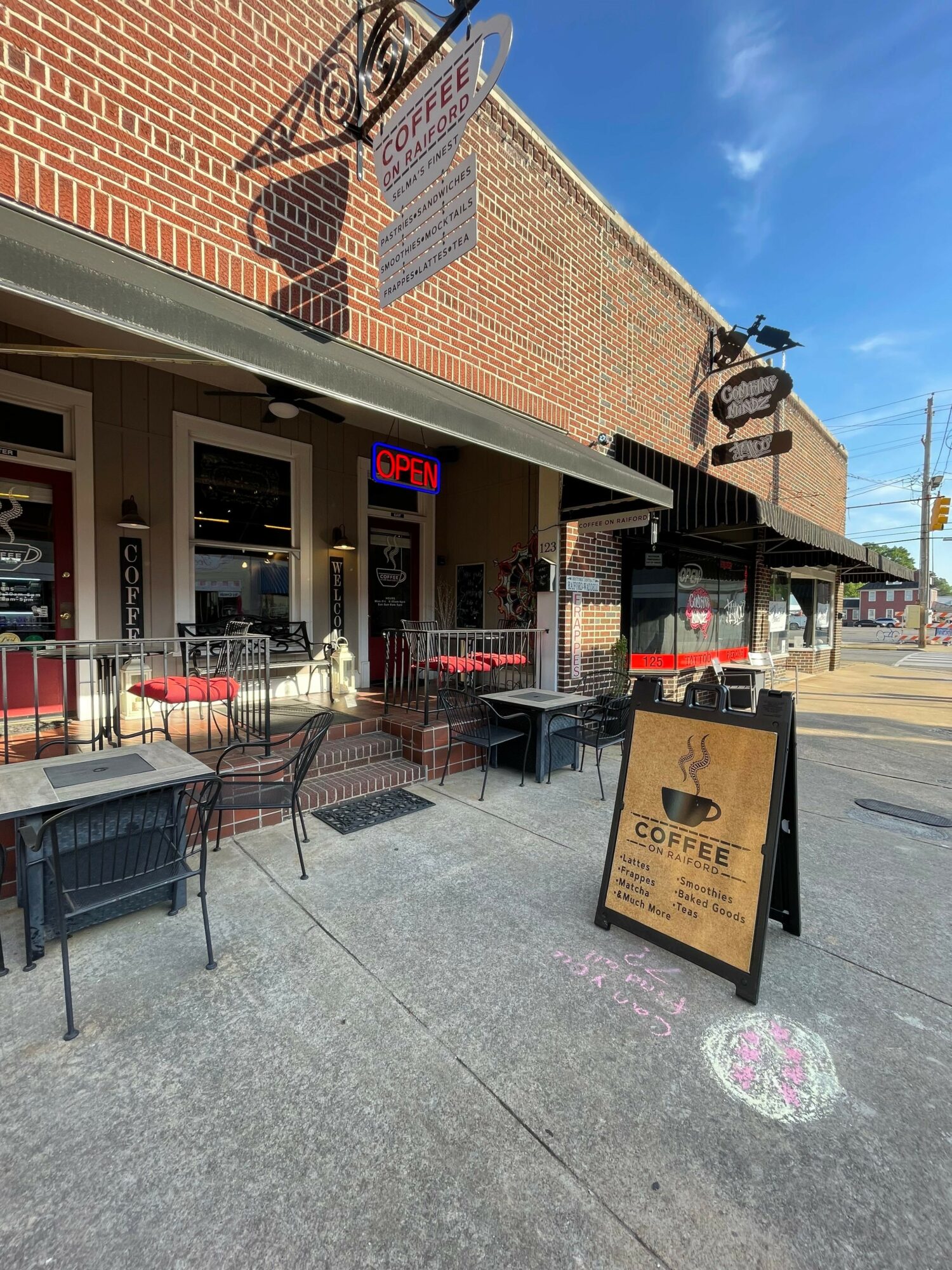 Street view of a small restaurant with outdoor seating, a signboard, and a brick building, under a blue sky.
