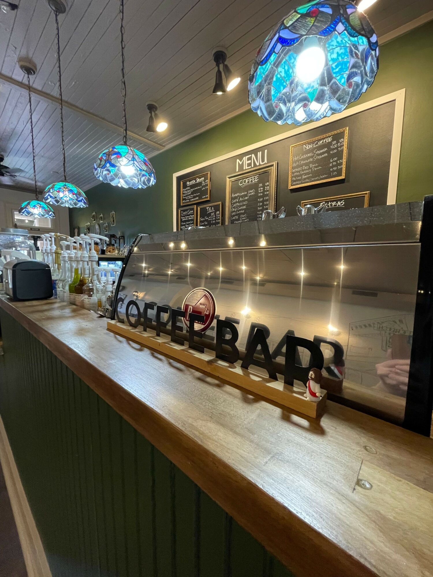 Bar counter with bottles and glasses, illuminated pendant lights, chalkboard menus, and a reflective surface behind the bar.