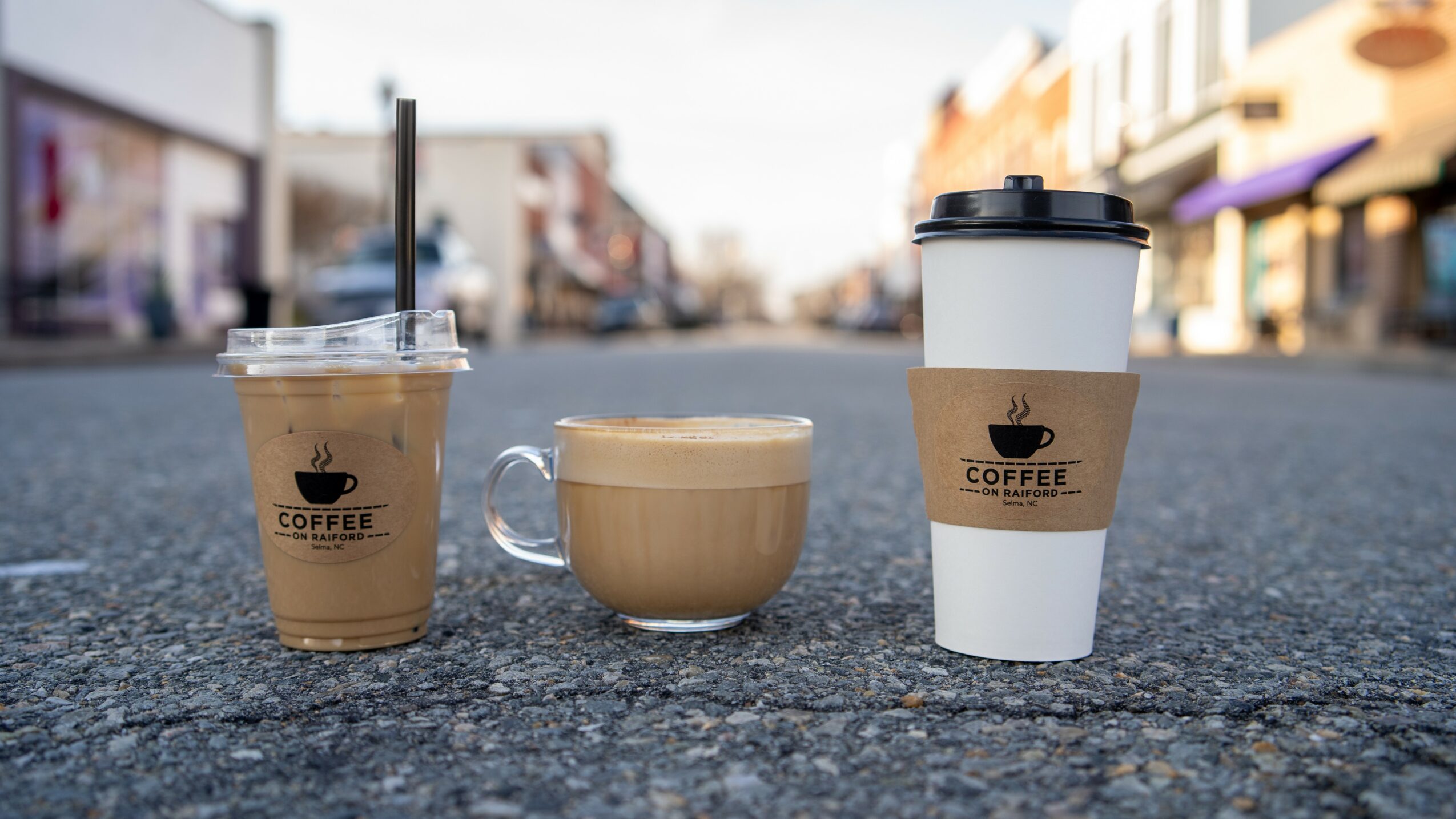 Three coffee drinks on a street: iced coffee, a latte, and a hot coffee in a paper cup, all with coffee logos.
