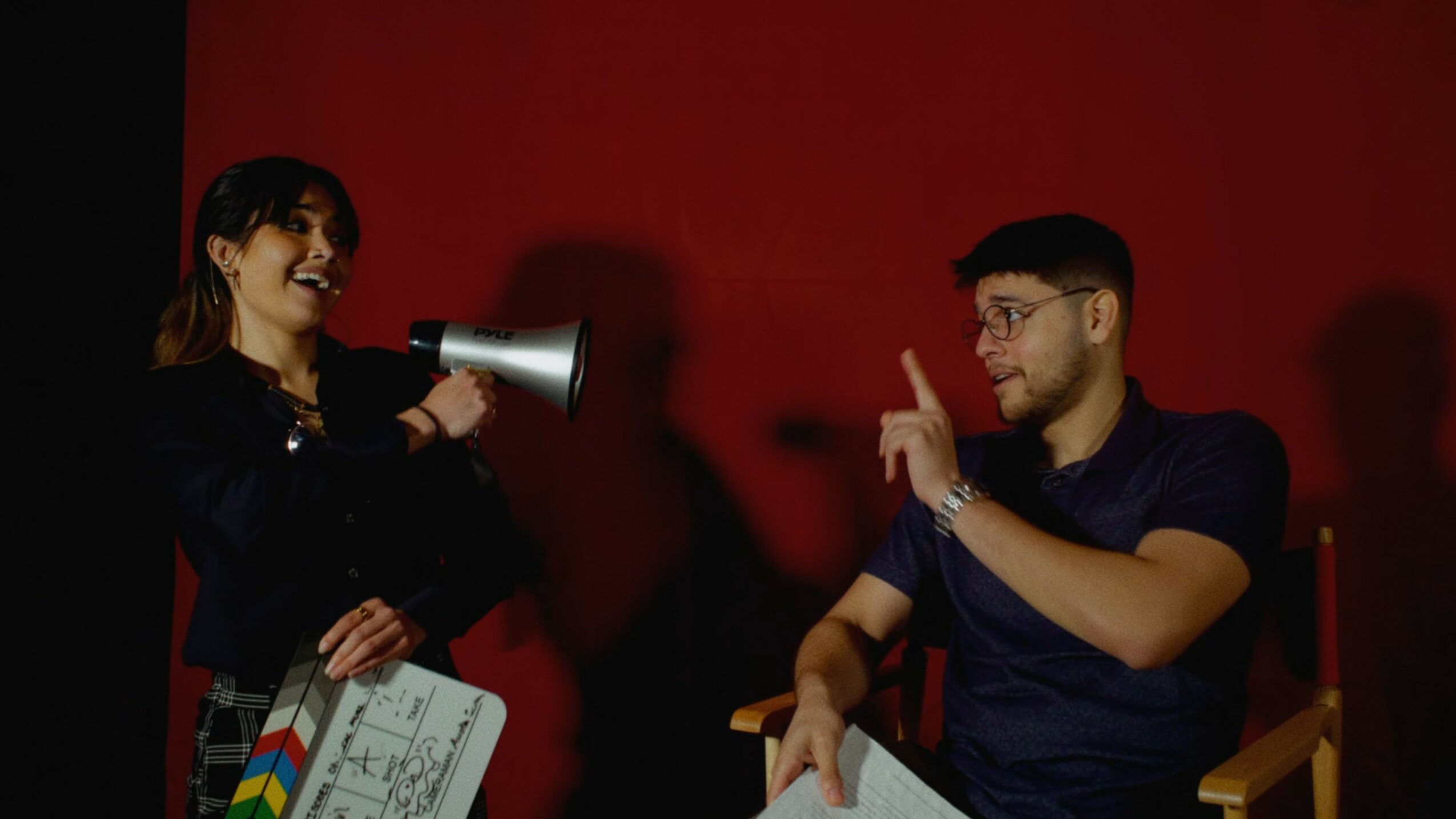 Two people engaged in a conversation, one holding a megaphone and a clapperboard, against a red background.