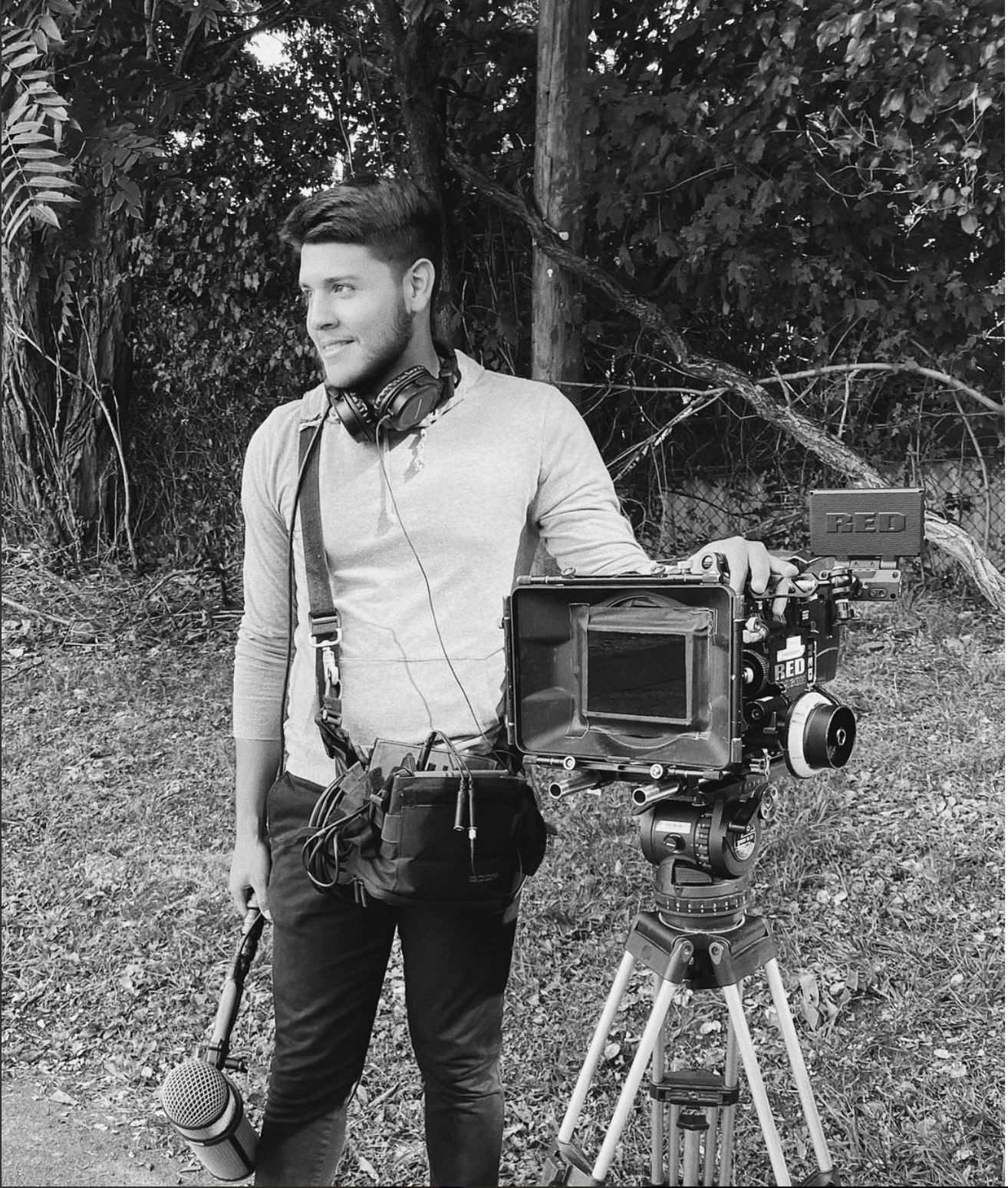 Young man with headphones standing outdoors next to a professional camera on a tripod, holding a microphone, surrounded by trees.