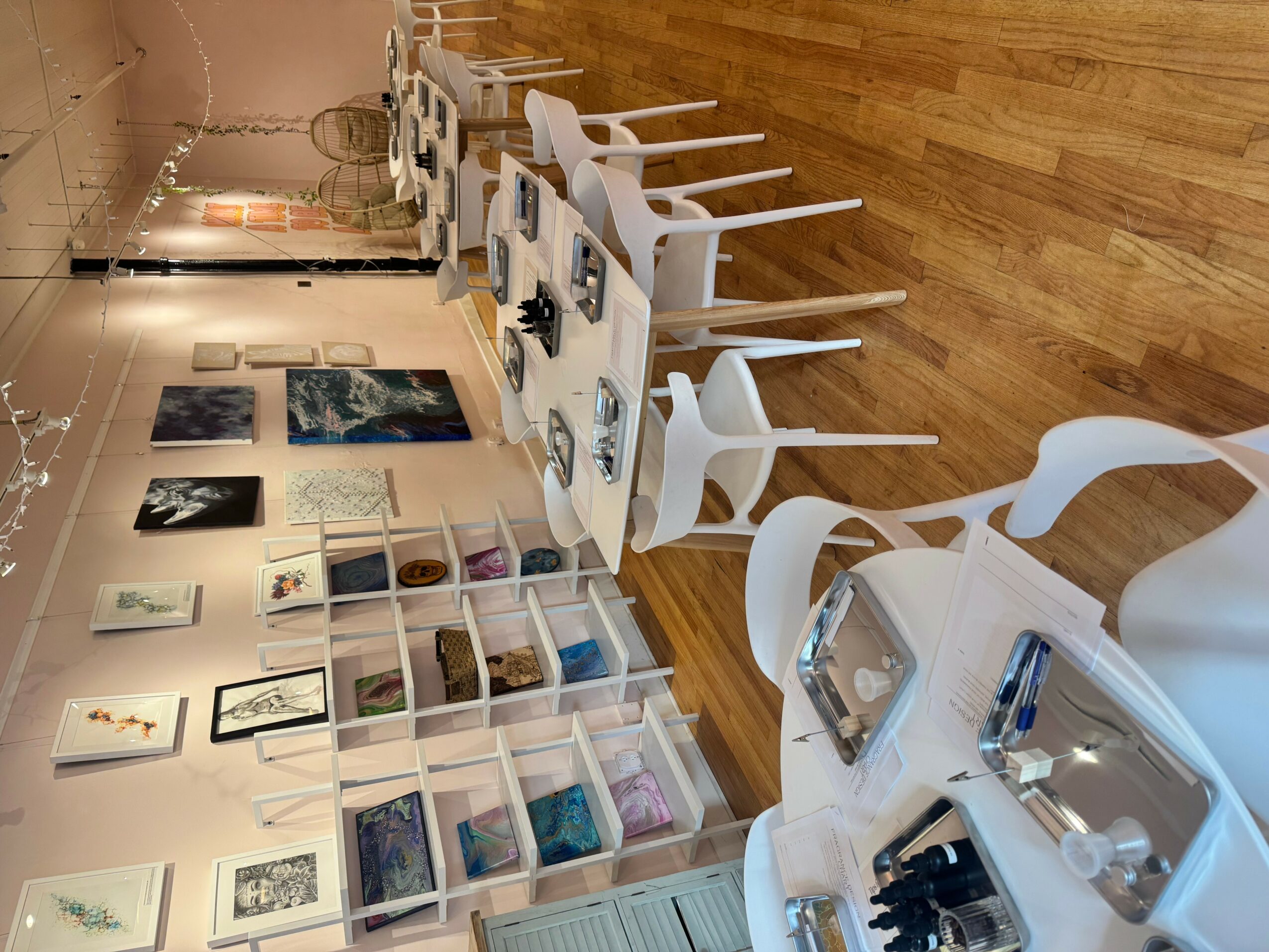 Gallery with framed artwork on wall, white chairs around tables, and display shelves with books and objects.