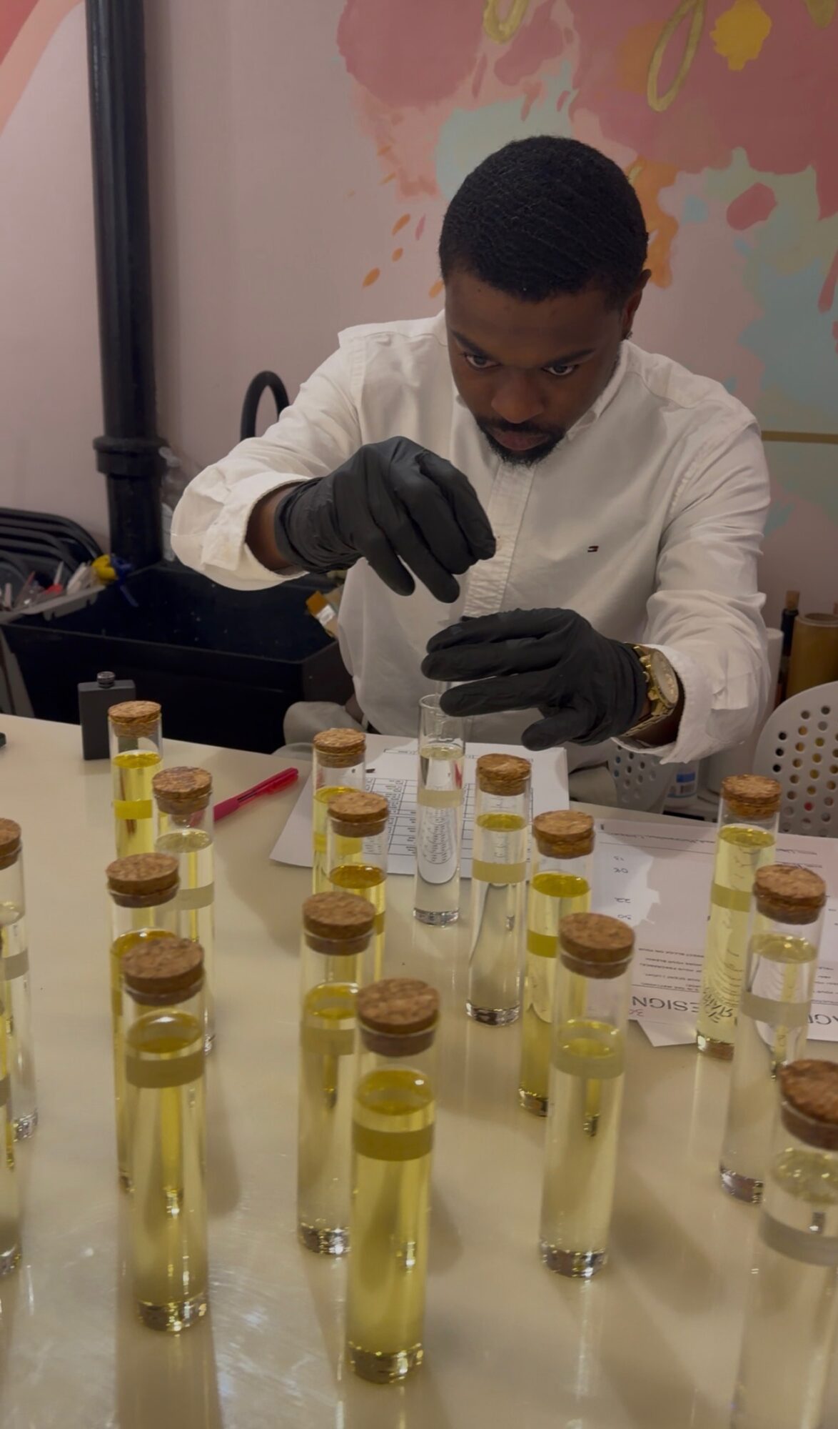 Man in white coat and black gloves examines small vials on table, colorful wall in background.