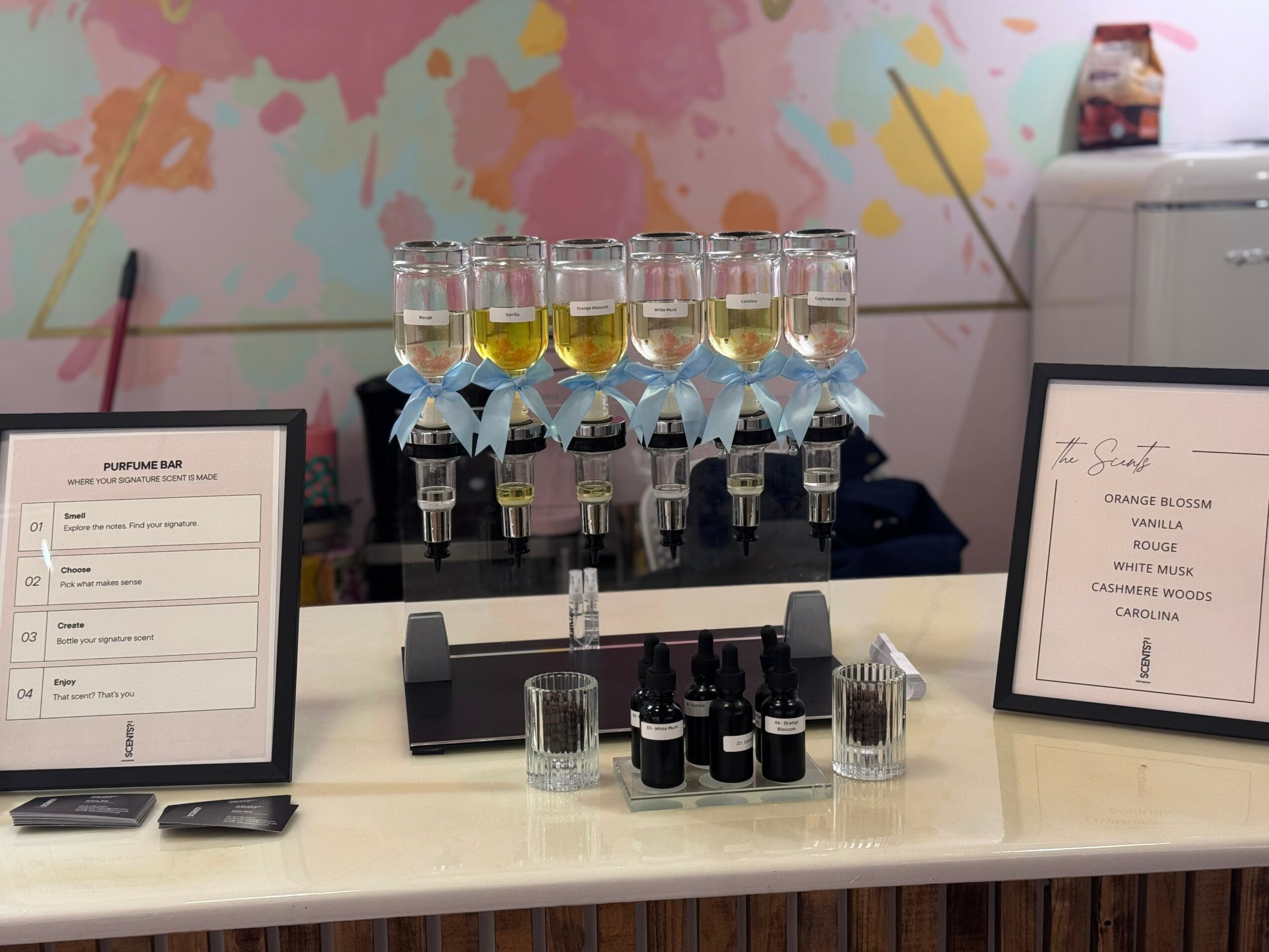 Display with glasses of champagne, bottles, and framed menus on a white table, colorful wall in background.