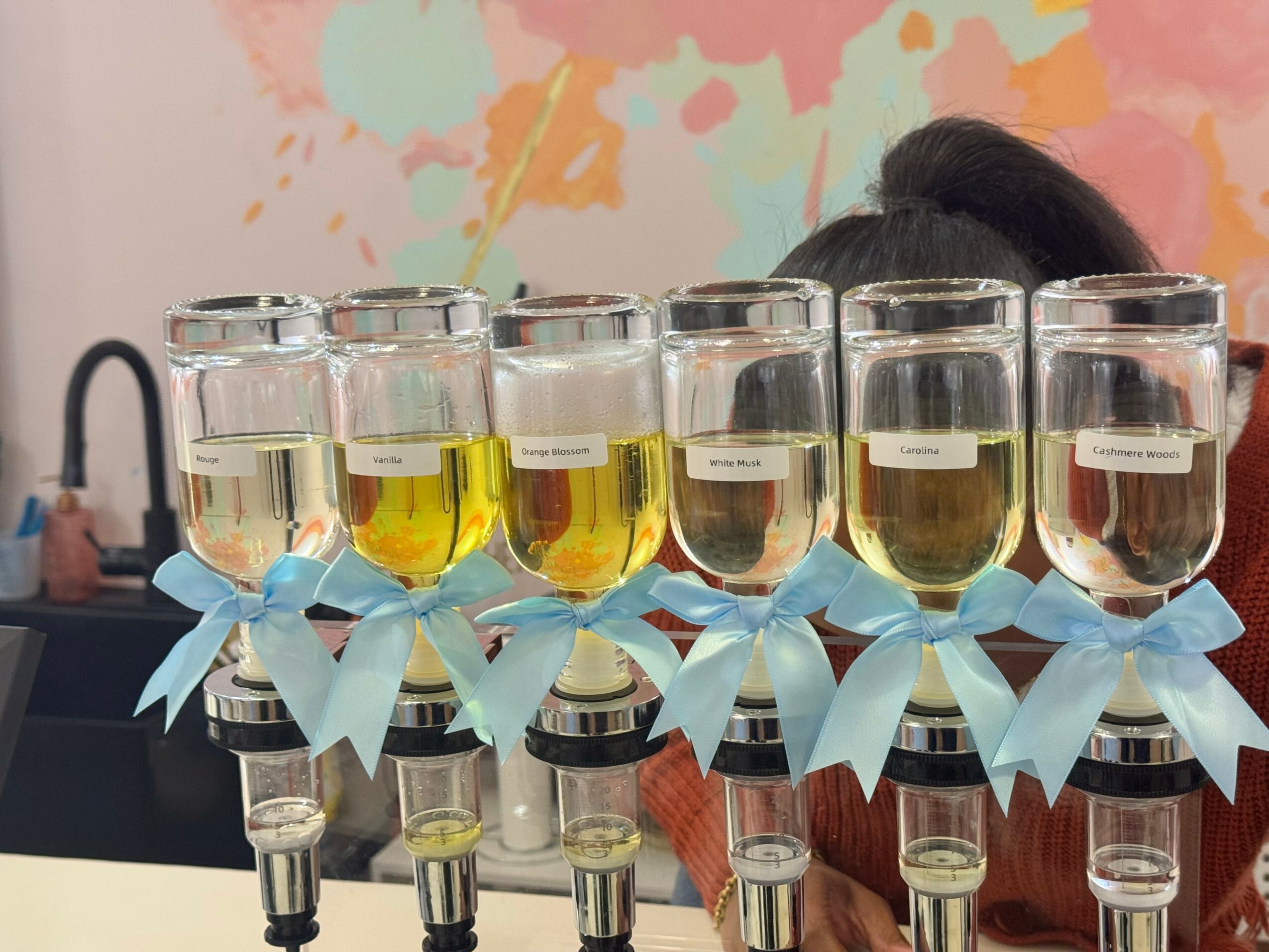 Six glass bottles with yellow liquid, decorated with blue ribbons, on a table, with a colorful wall in the background.
