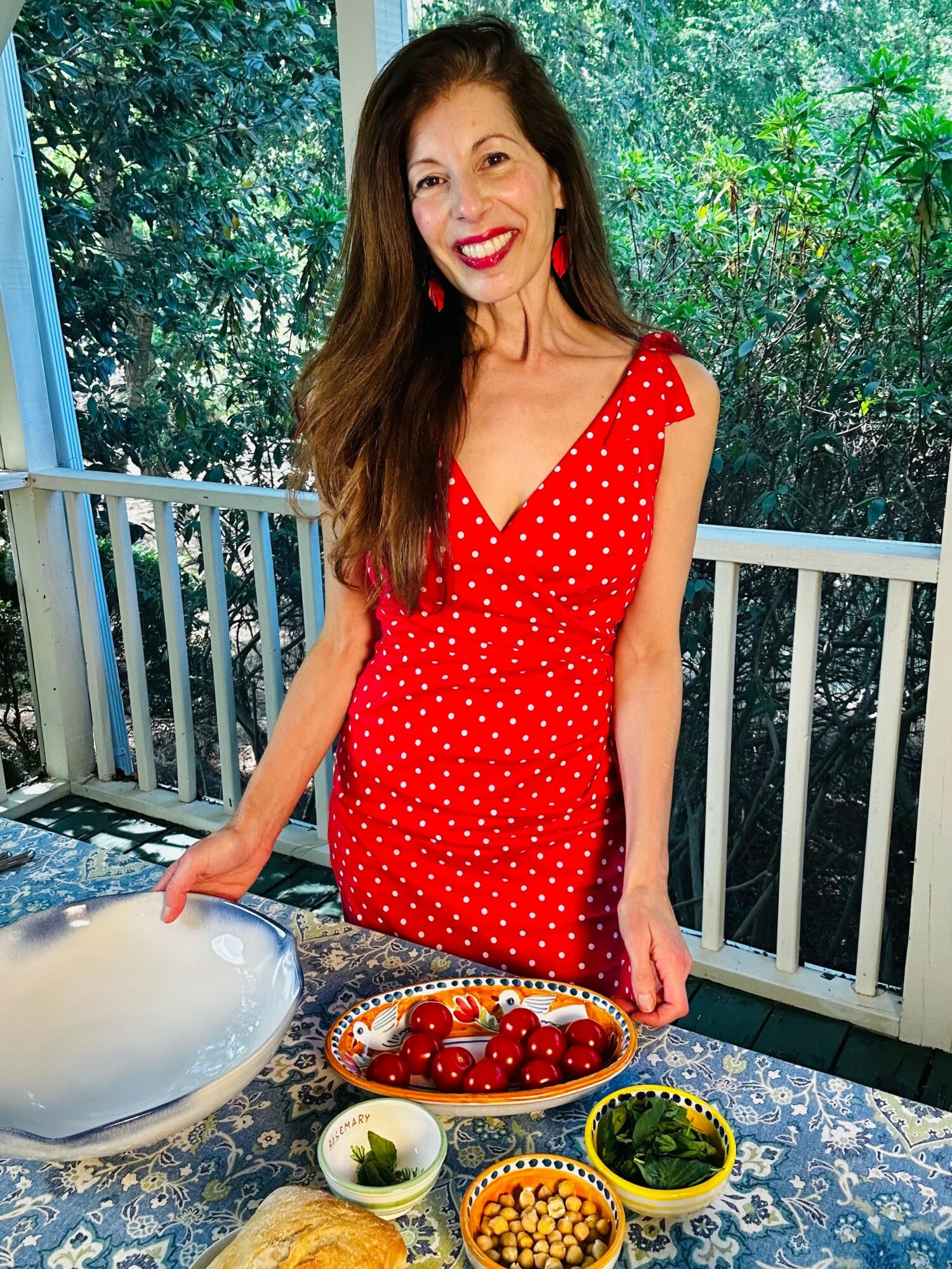 Woman in red polka dot dress smiling on balcony with trees in background, standing at table with bowls of food.