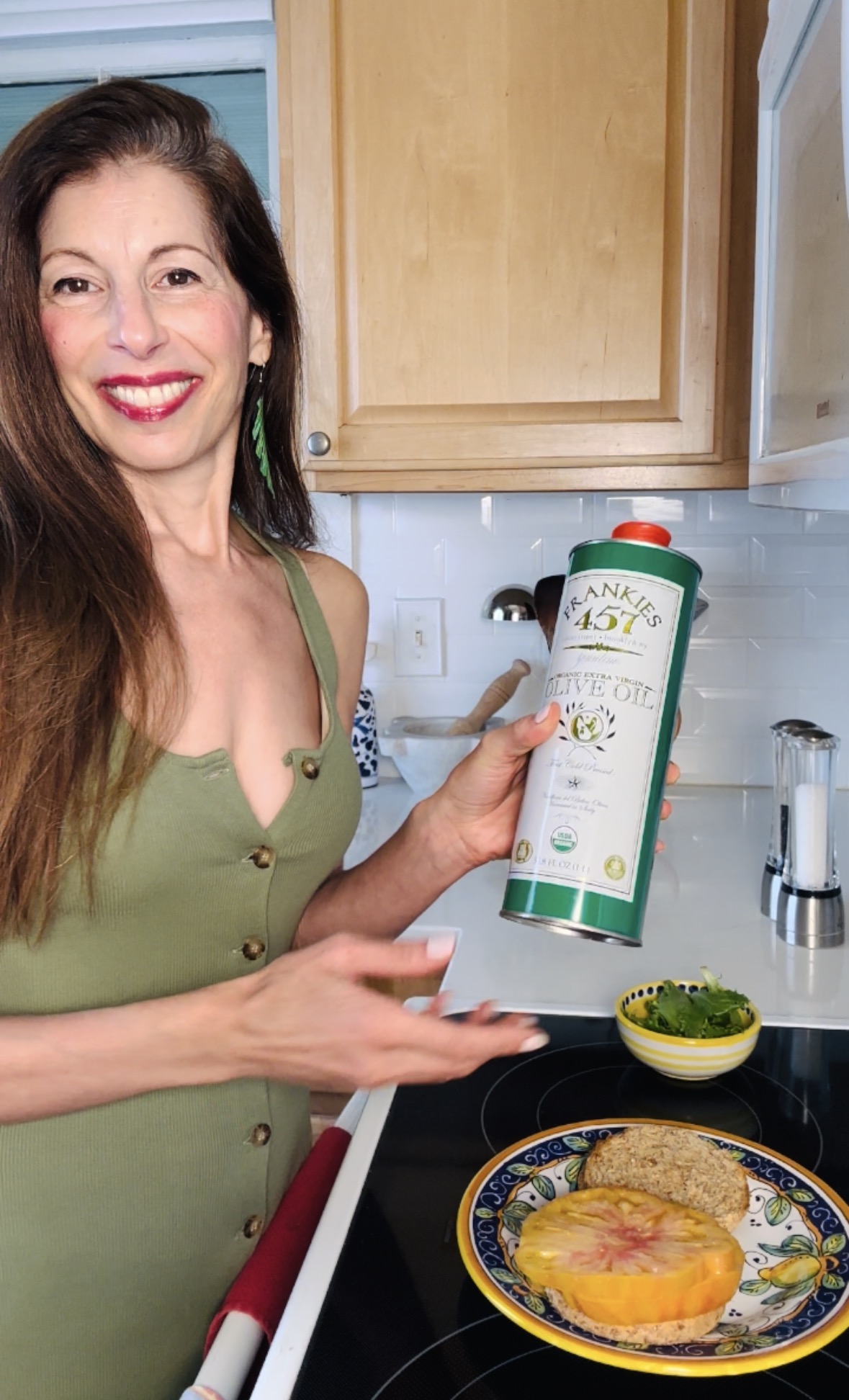 Woman smiling in kitchen holding a container of plant-based food, with a plate of food and a bowl nearby.