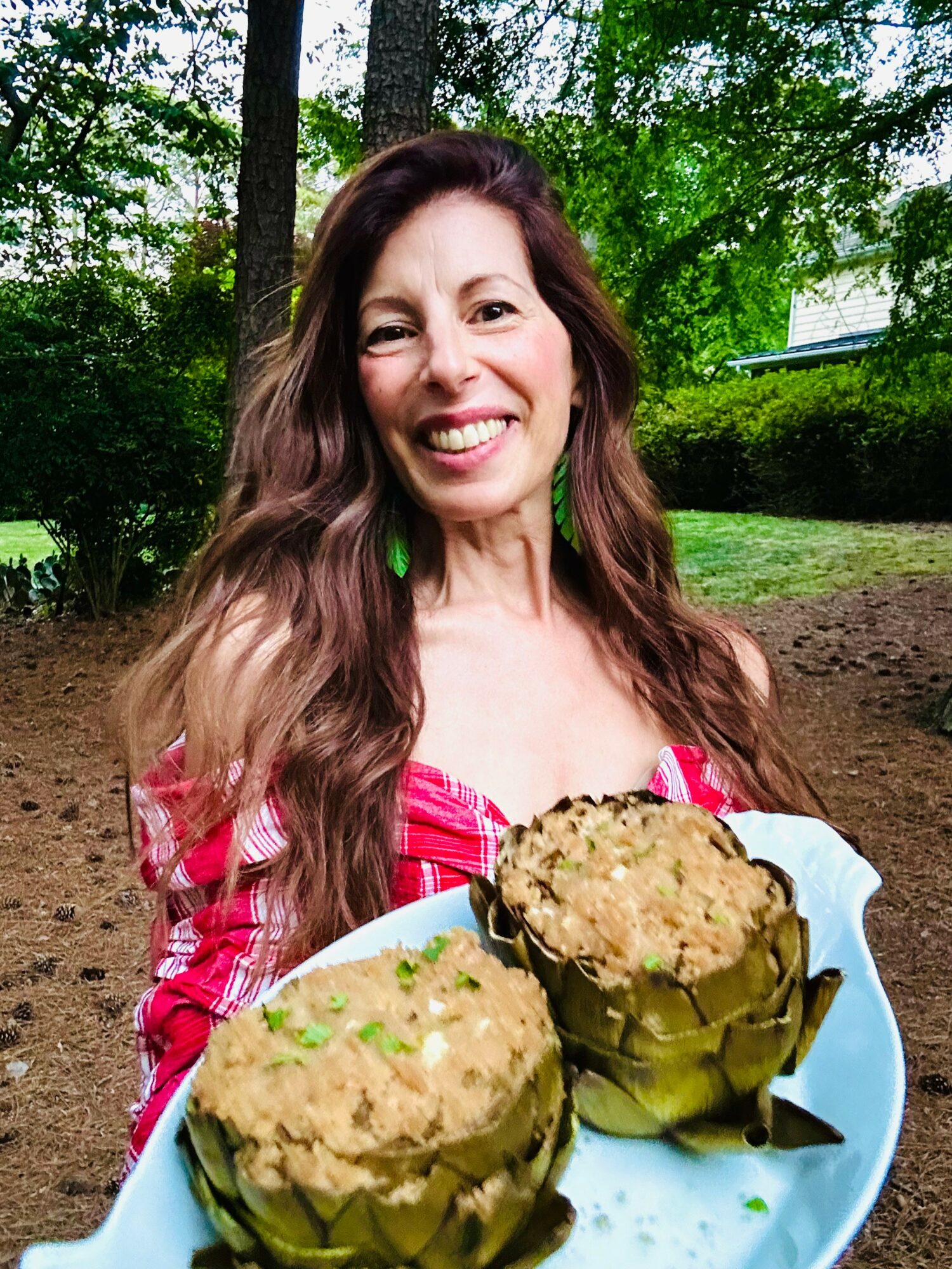 Woman smiling outdoors holding a tray with two baked dishes in green ceramic bowls, surrounded by trees and greenery.