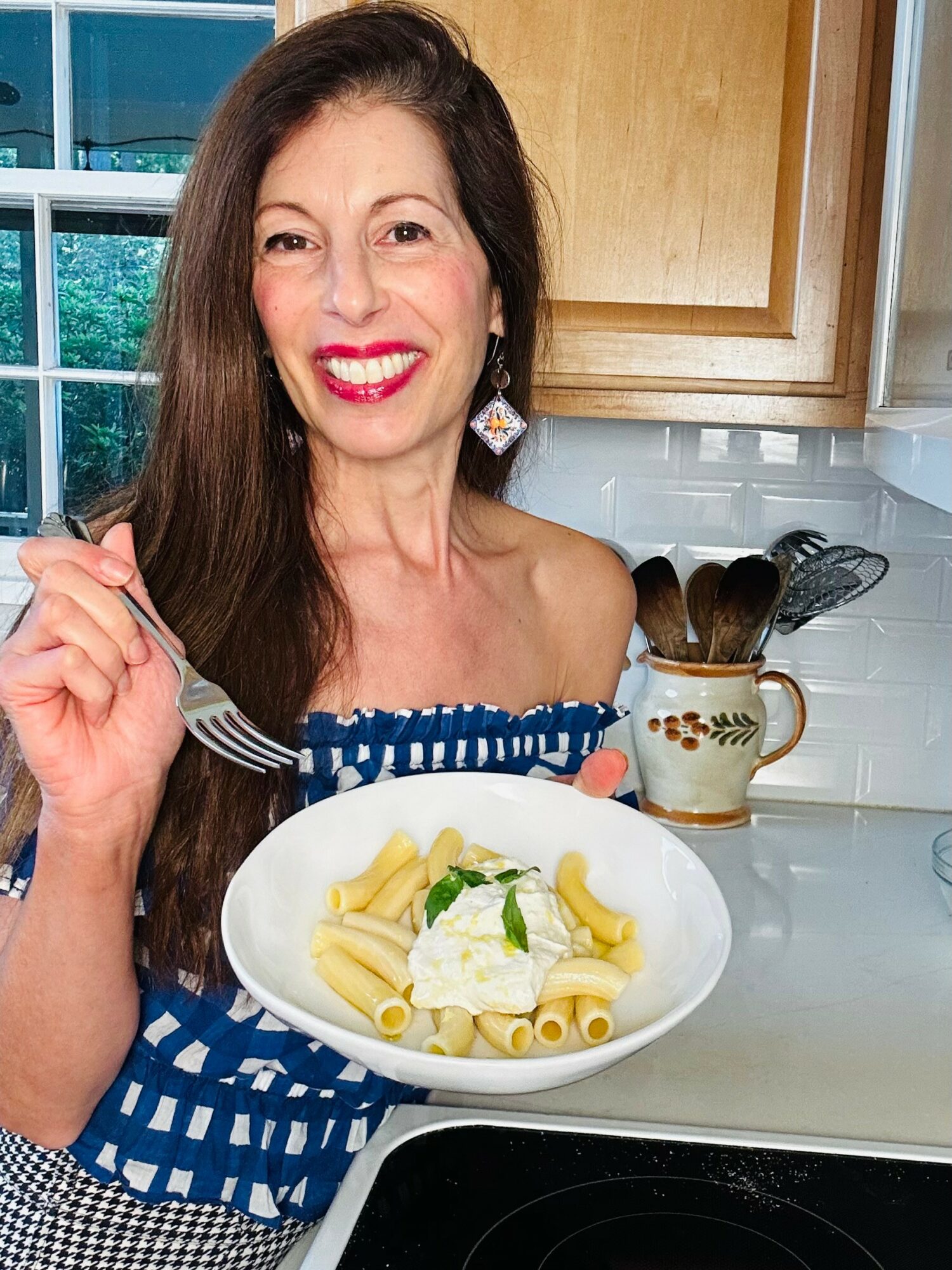 Woman smiling holding a plate of pasta in a kitchen, wearing a blue and white dress, with wooden cabinets in the background.