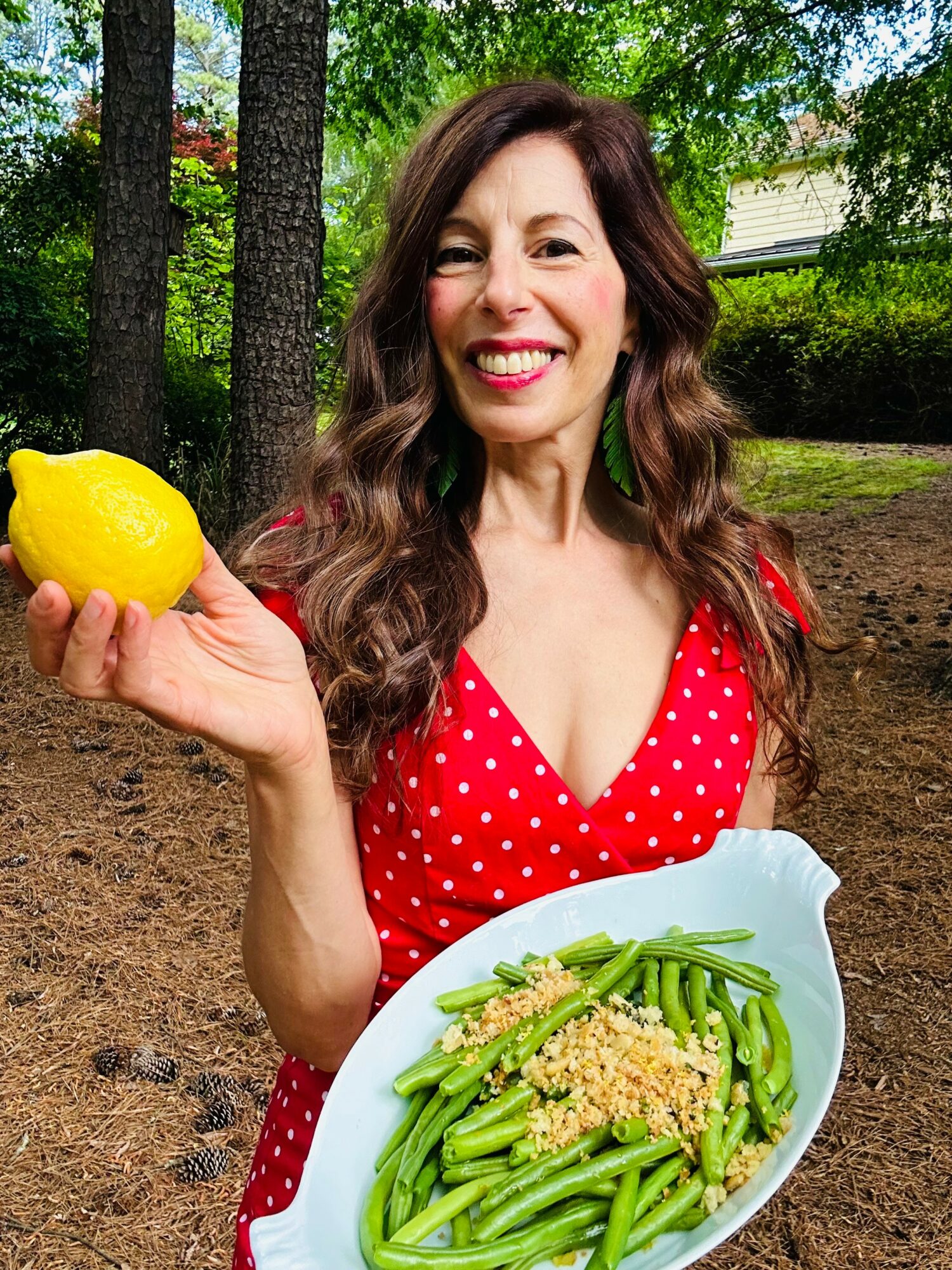 Woman with long brown hair smiling outdoors, holding a lemon, wearing a red polka dot dress, with a plate of green beans and grains.