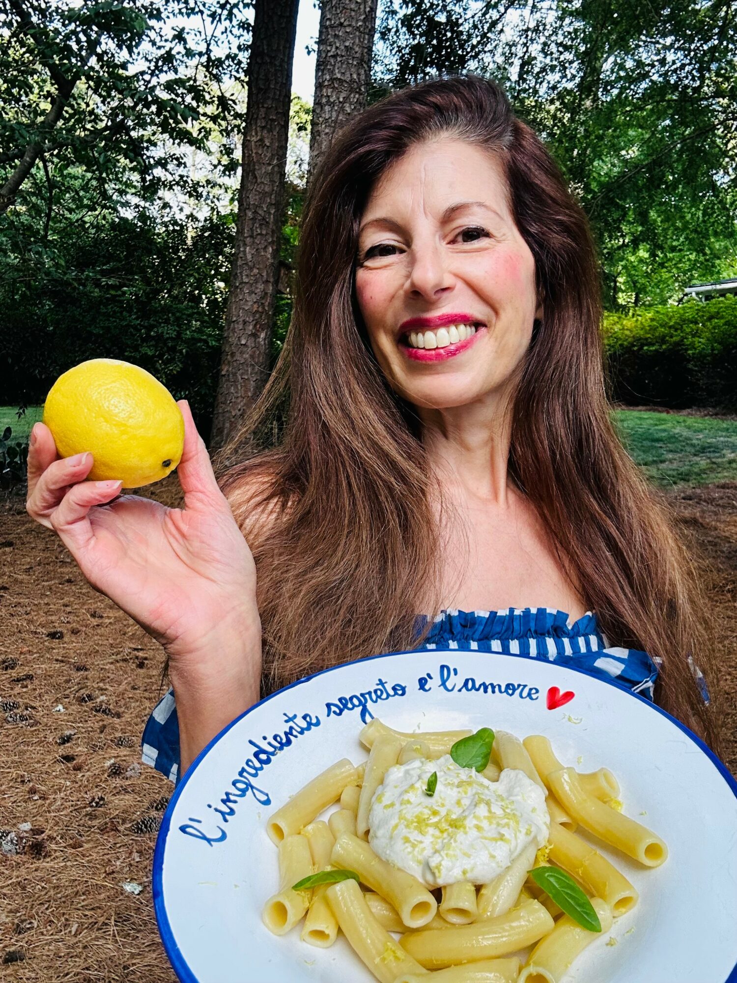 Woman smiling outdoors holding a lemon and a plate of pasta with cheese and green garnish.