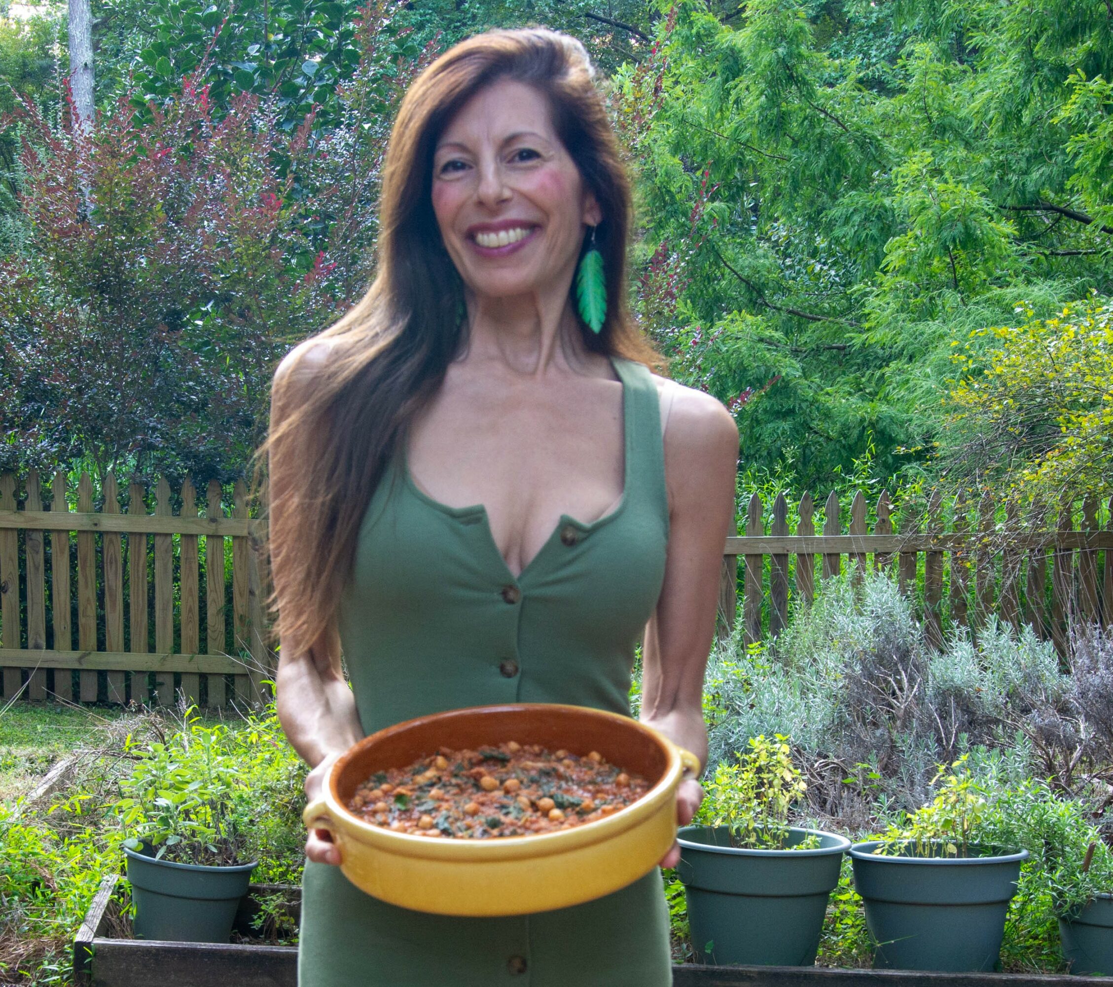 Woman with long hair smiling outdoors, holding a dish, surrounded by plants and trees.