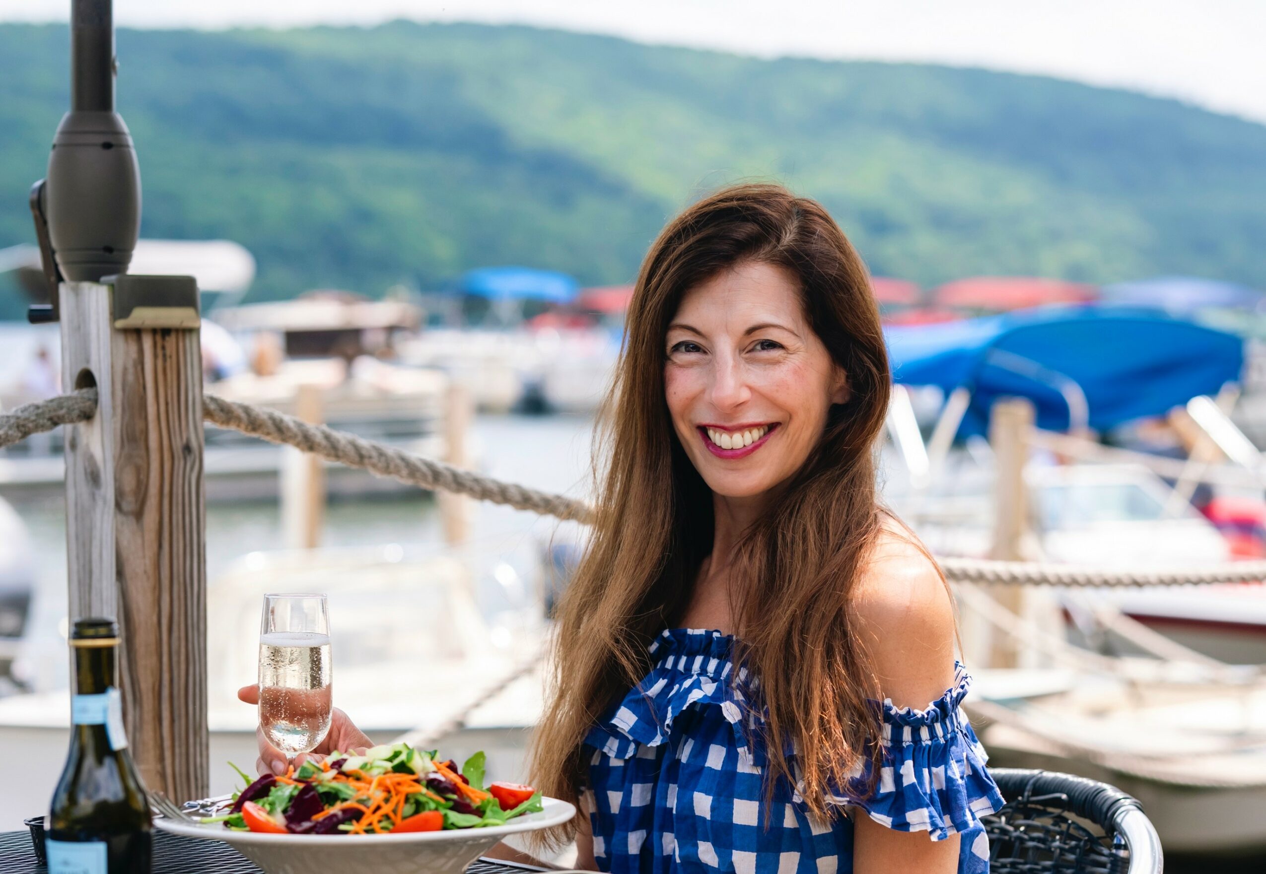 Woman with long hair smiling, holding a glass of wine, sitting outdoors near a marina with boats and umbrellas in background.
