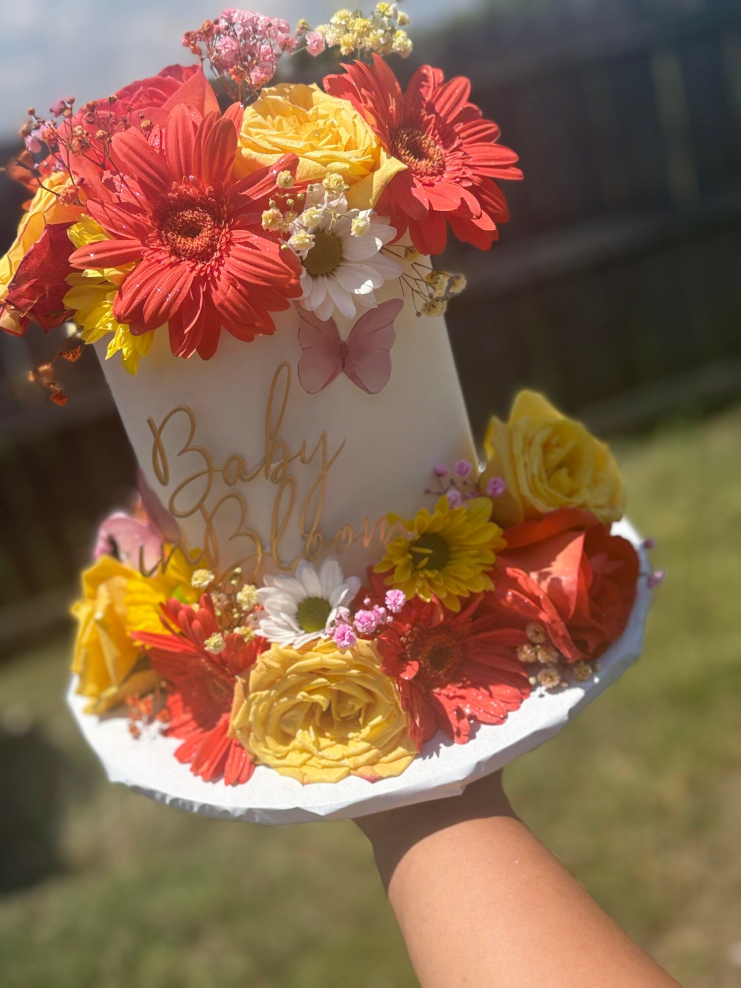 Colorful flower arrangement with red, yellow, and pink flowers in a white container, outdoors on a sunny day.