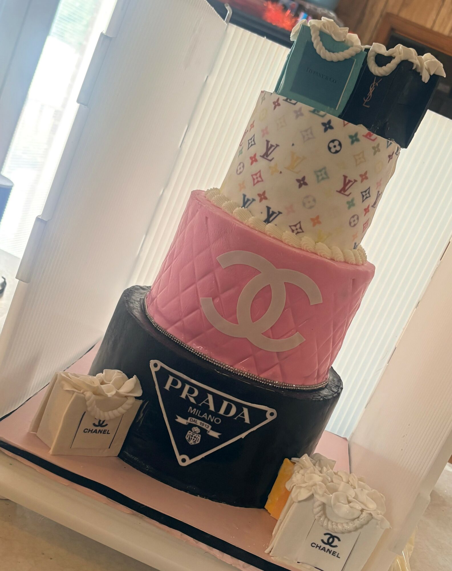 Four-tiered cake with designer logos and patterns, decorated with flowers, on a white tray in a room with natural light.