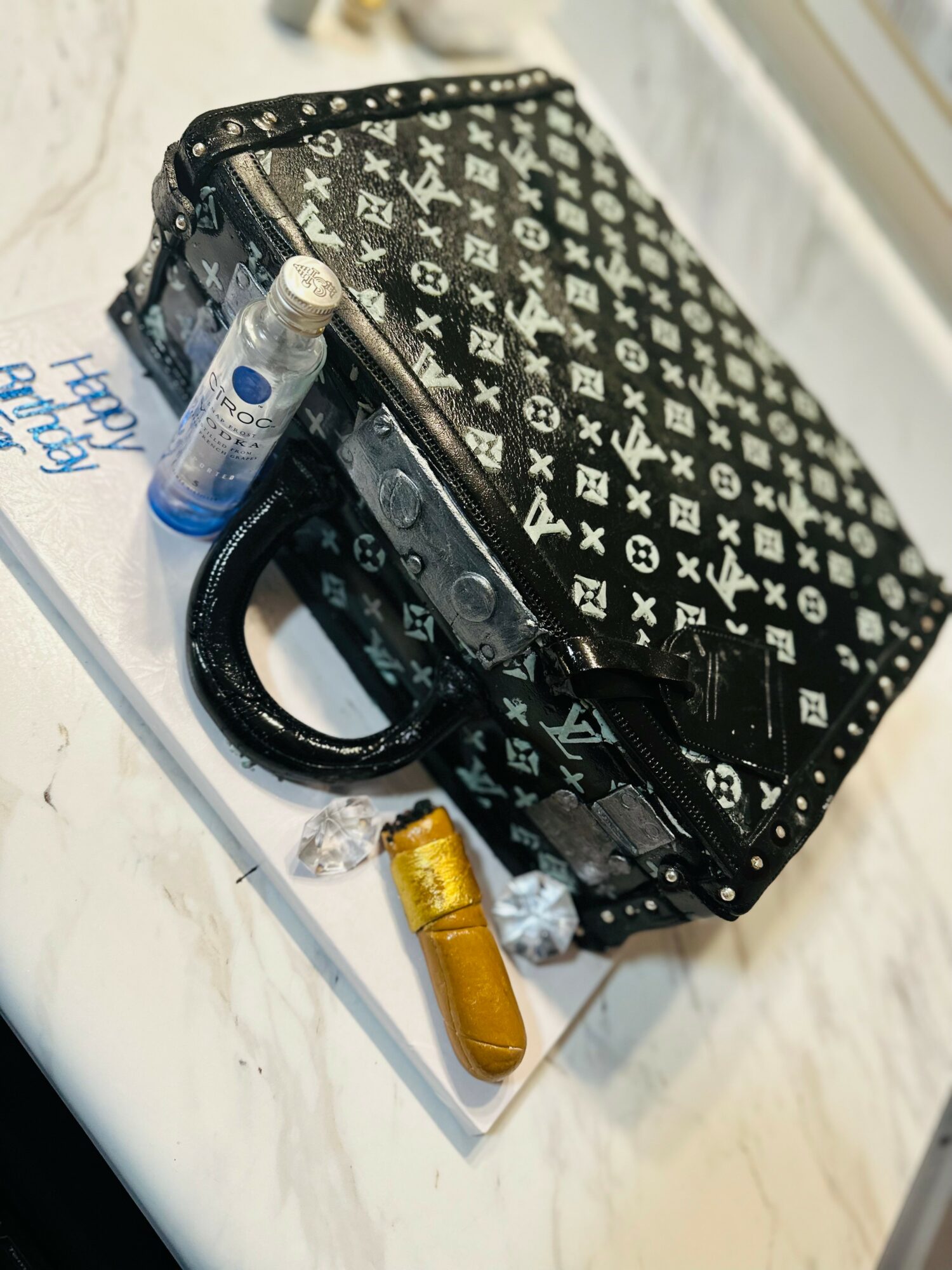 Black patterned handbag with a handle, placed on a white surface with small items nearby, including a small bottle and a lighter.