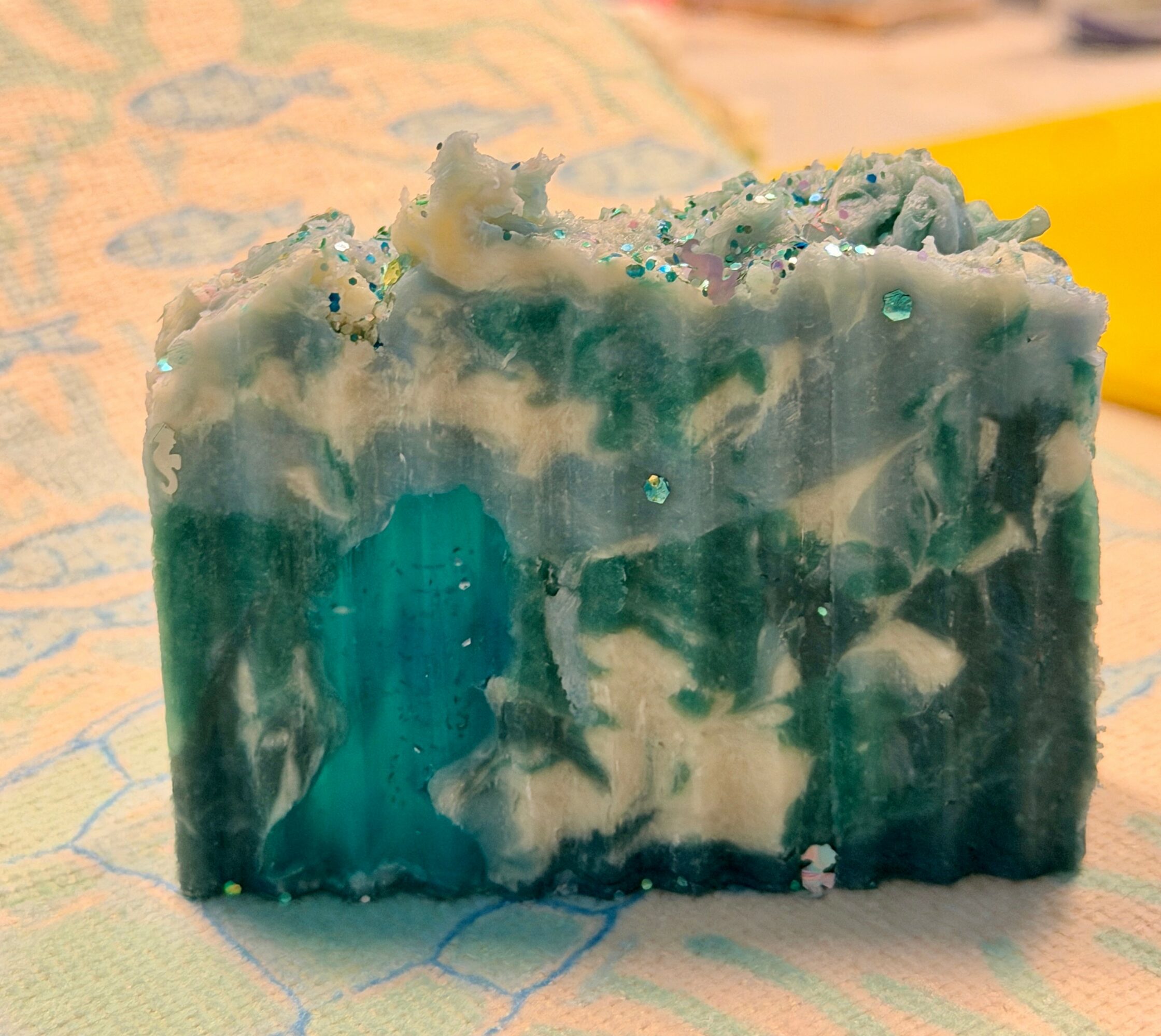 A bar of soap with blue, white, and green marbled colors, placed on a surface with a patterned background.