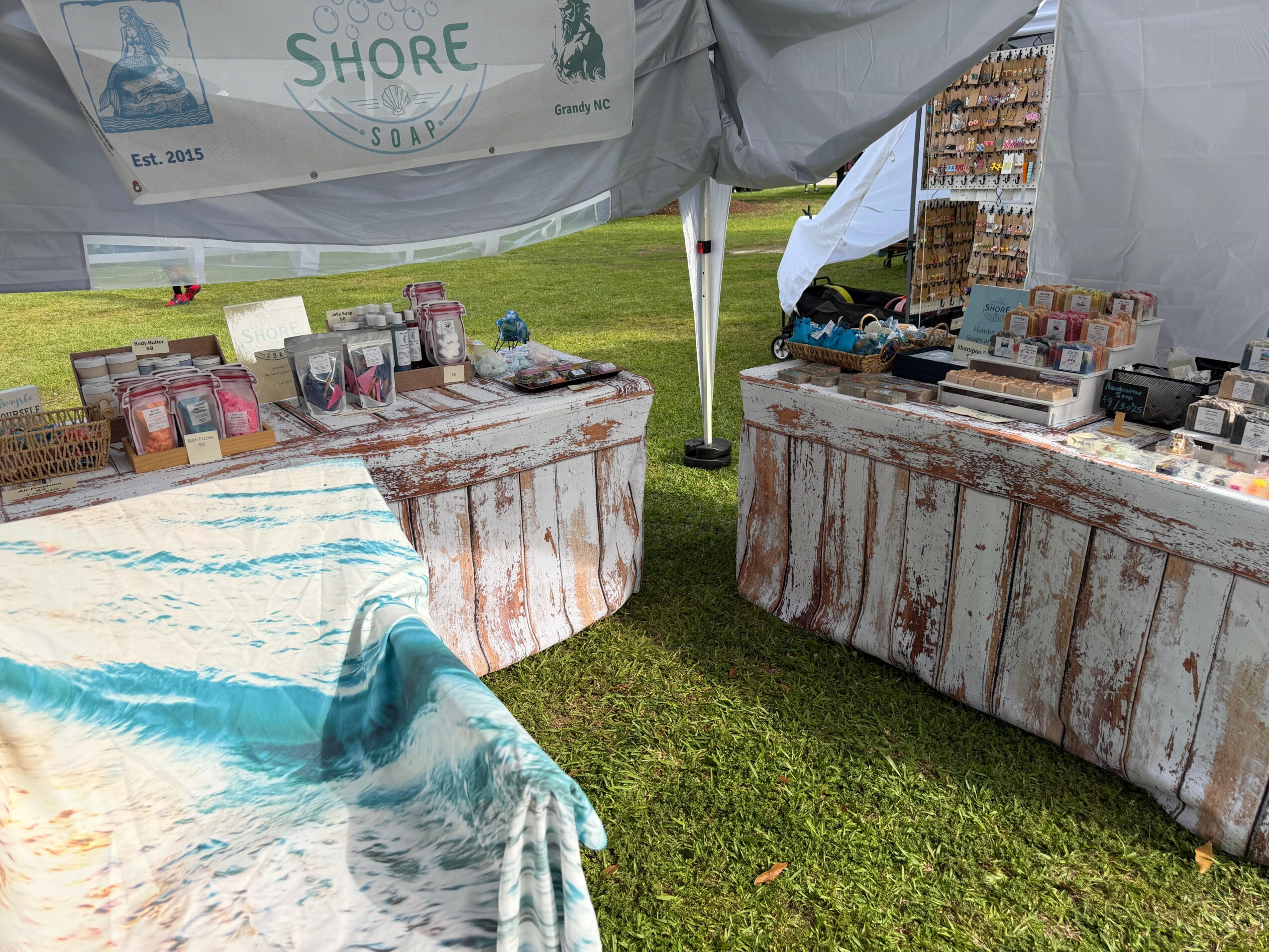Outdoor market stall with tables displaying various products, set on grass under a tent.