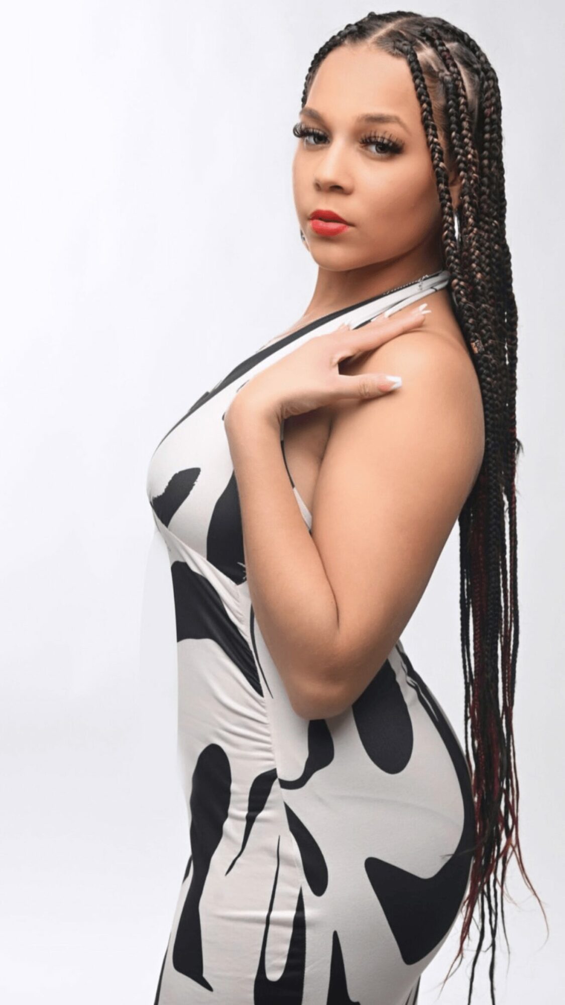 Young woman with braided hair in a black and white patterned dress, posing with hand near shoulder, against a white background.