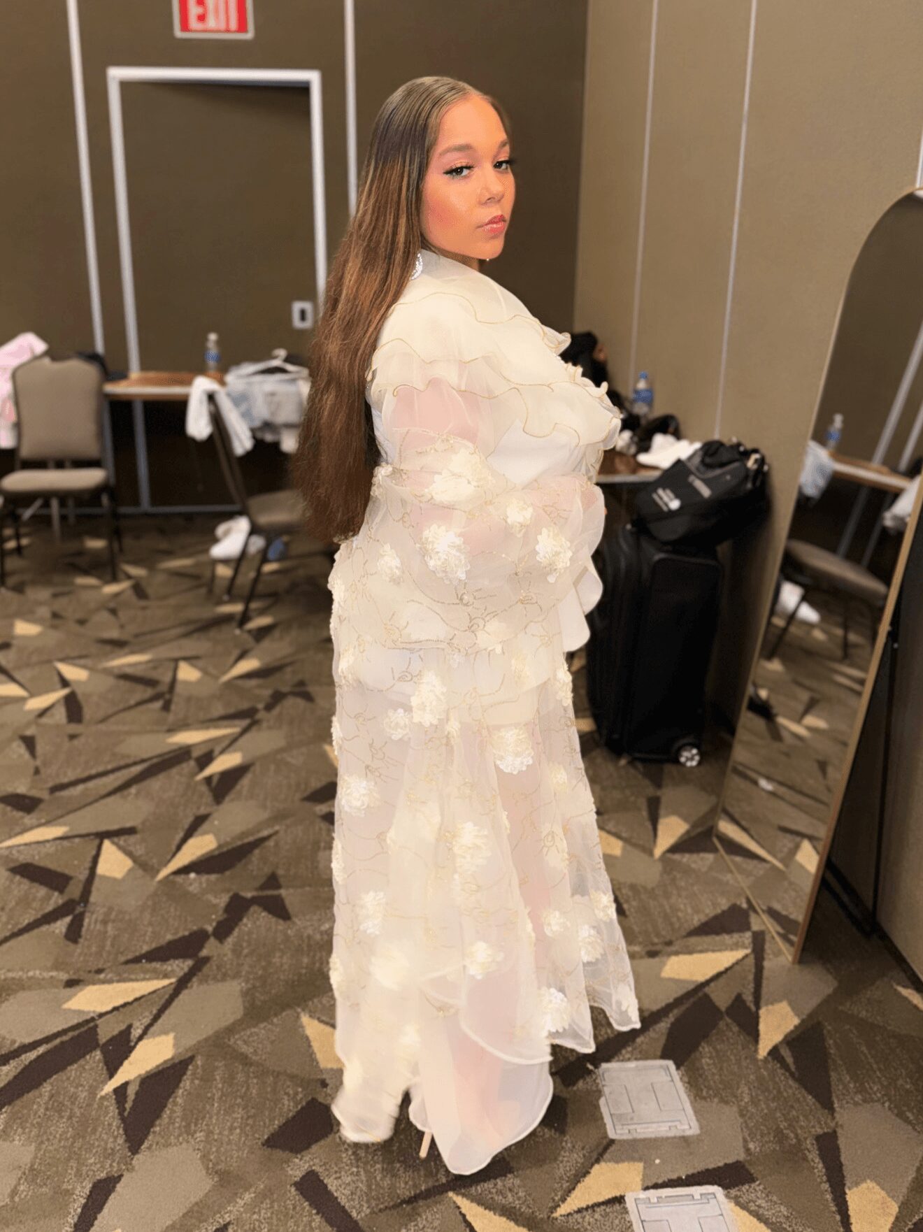 Woman with long hair in a white dress standing in a room with chairs and tables, facing sideways.