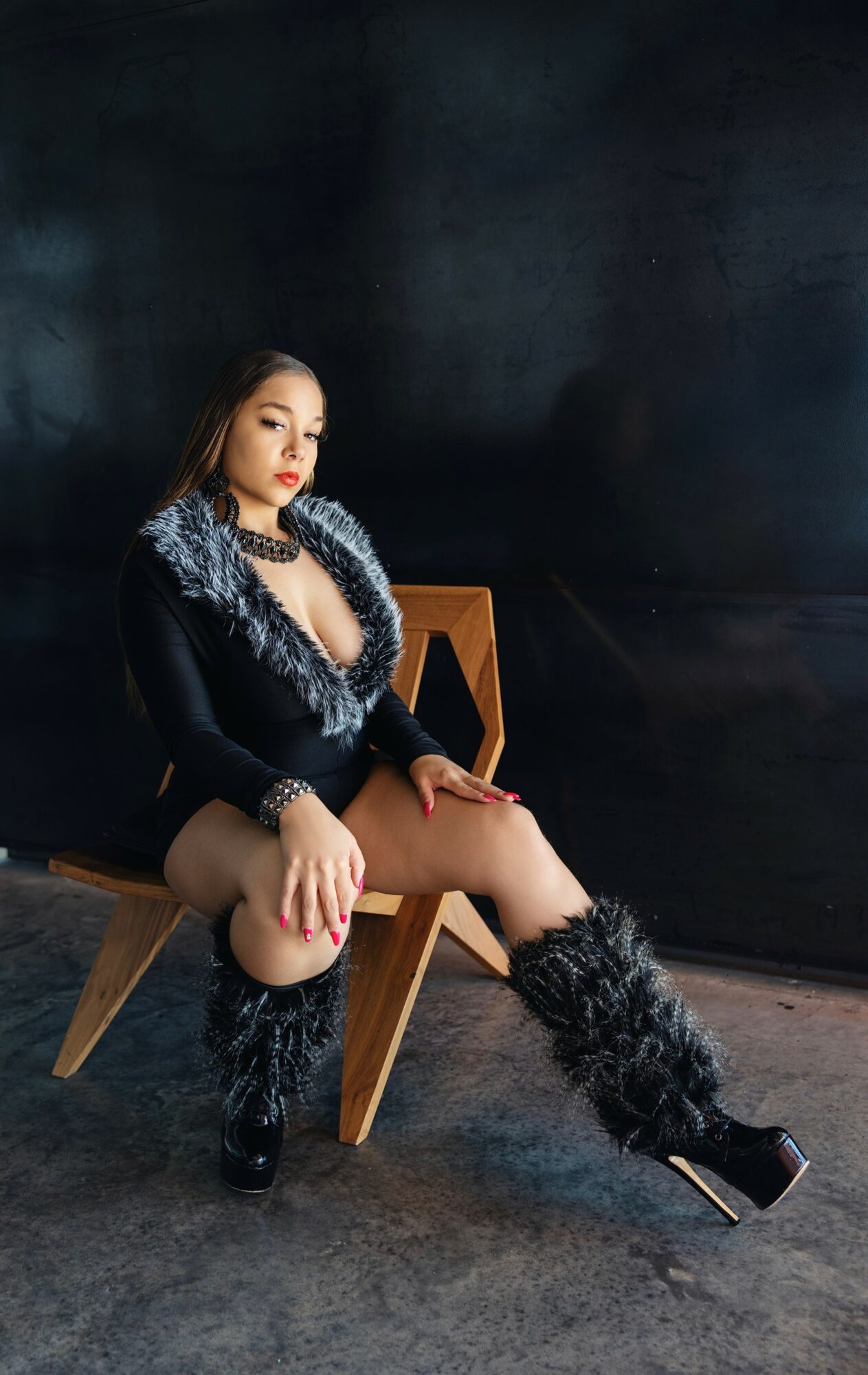 Woman sitting on a wooden chair, wearing black and silver outfit with furry boots, against a dark background.