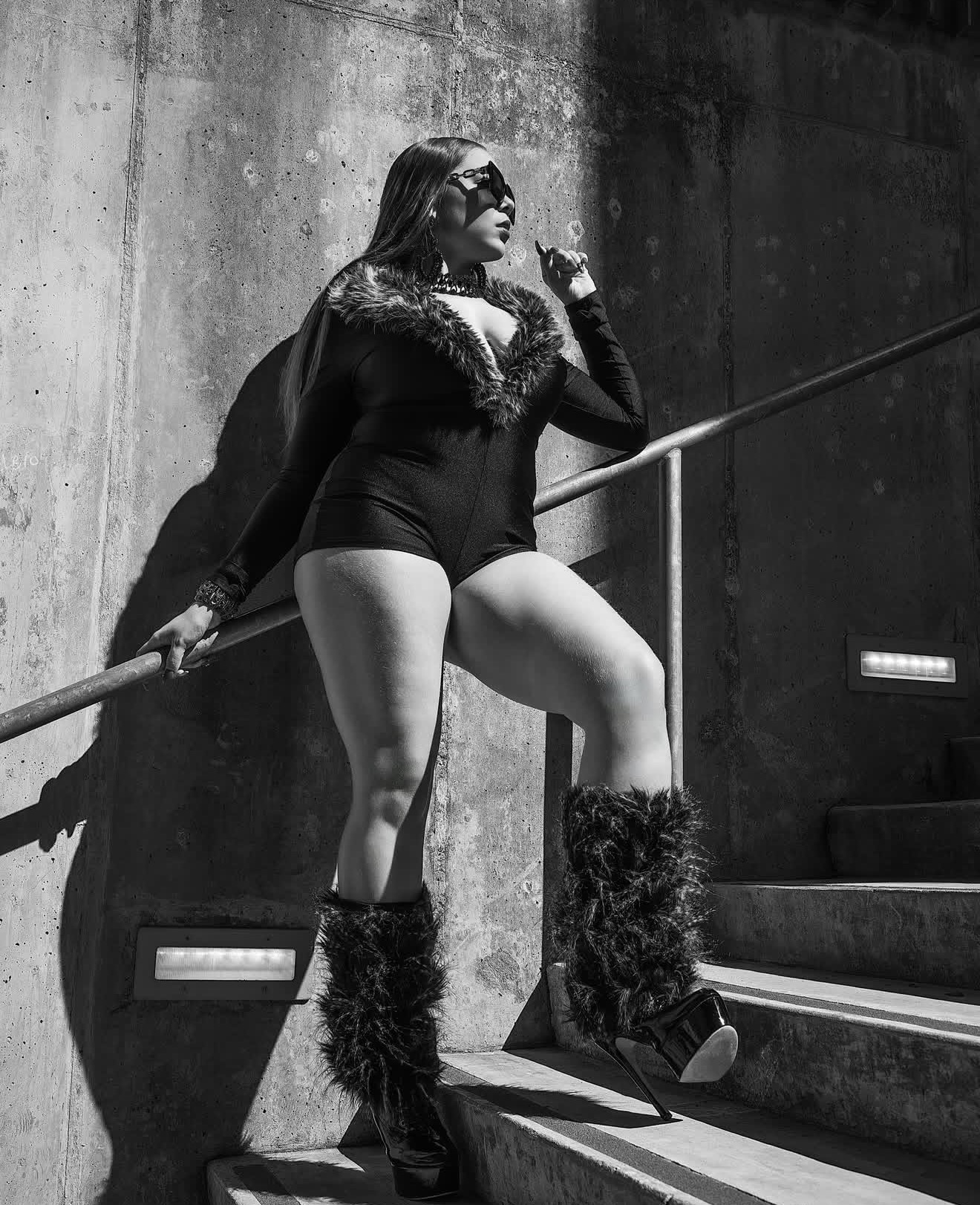 Woman in black outfit with fur boots standing on stairs, leaning against a wall, holding a railing, in black and white.