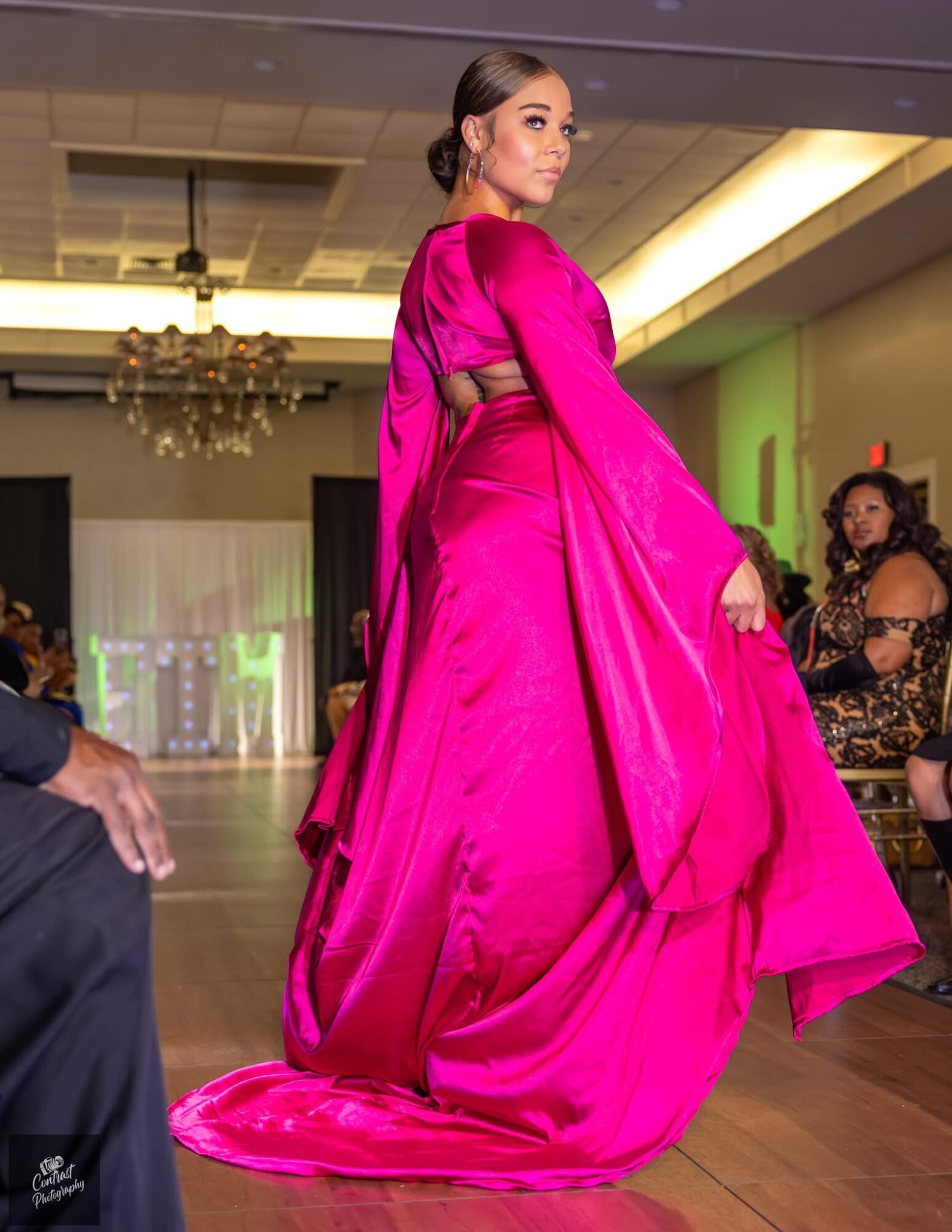 Woman in a bright pink gown walking on a runway with an audience and chandelier in background.