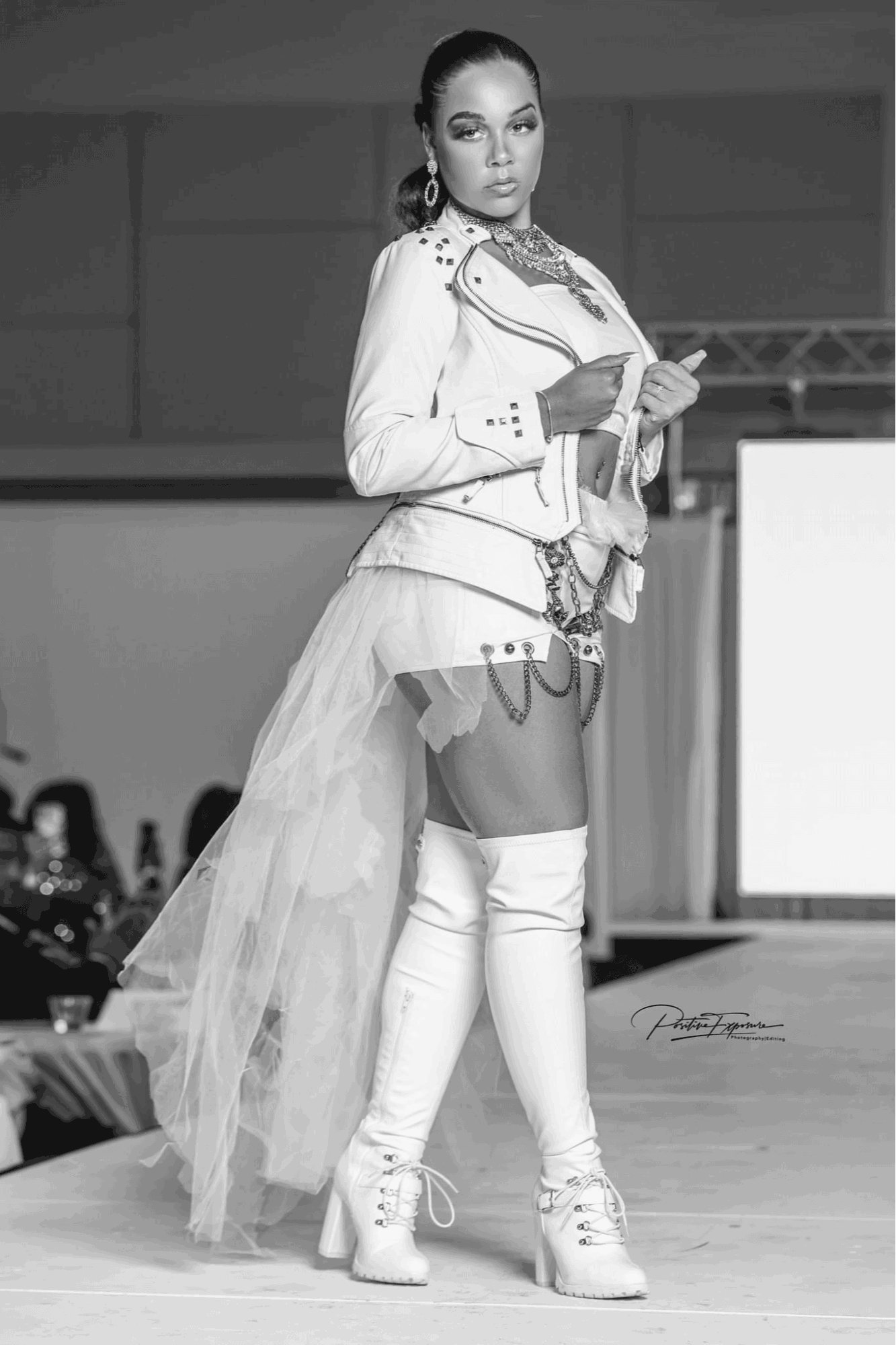 Woman in a jacket and thigh-high boots on runway, holding a paper, with audience in background.