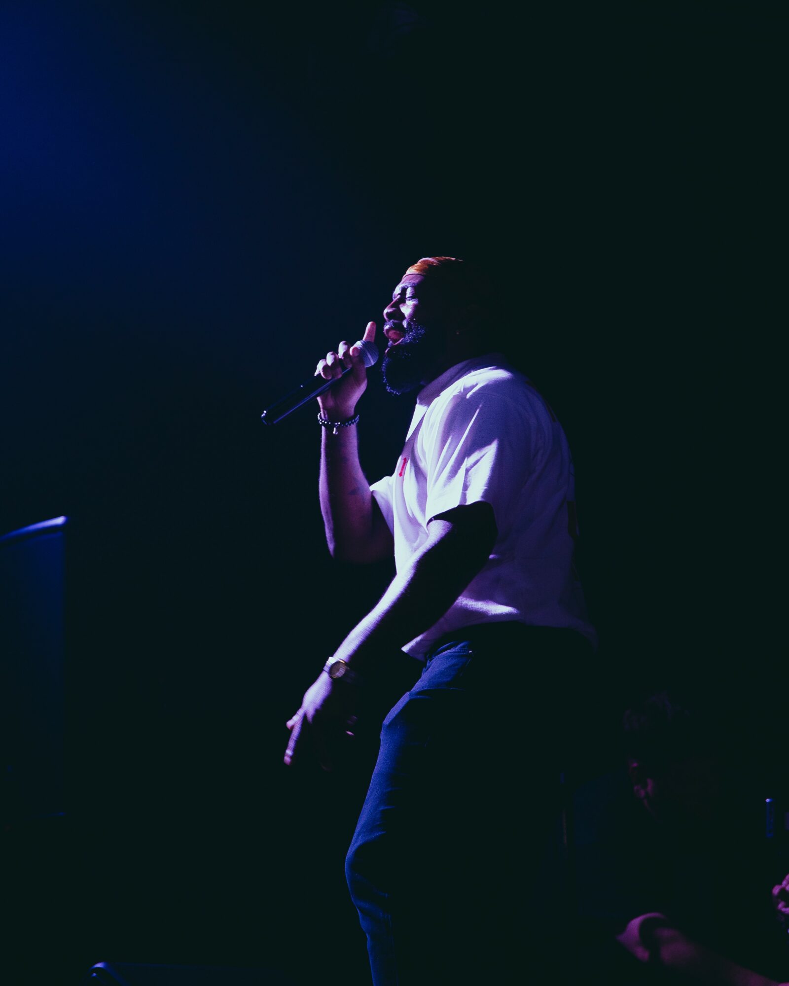 Person holding a microphone on stage, wearing a white shirt and dark pants, with a dark background.