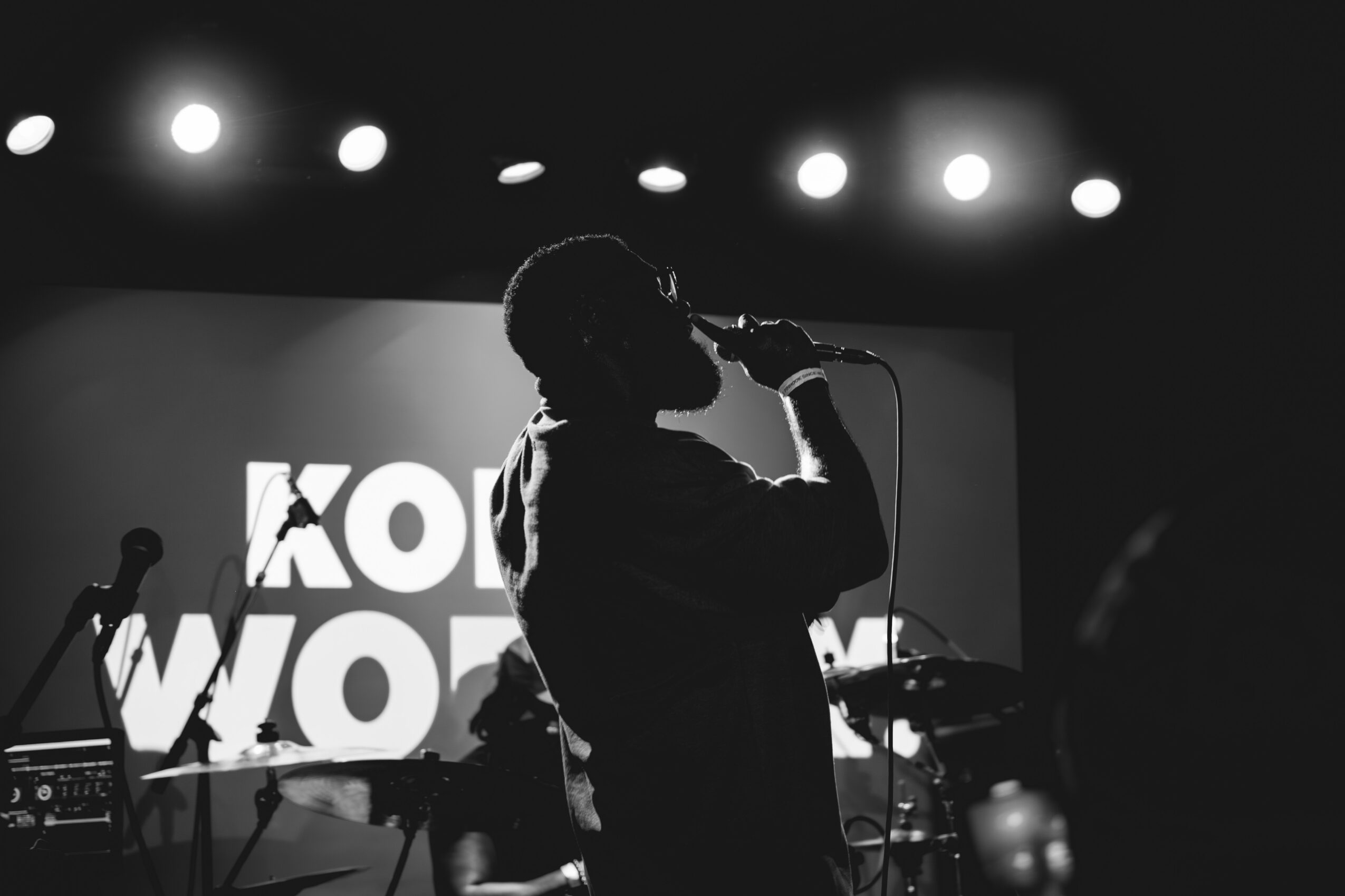 Silhouette of a person singing into a microphone on stage with bright lights and a screen in the background.