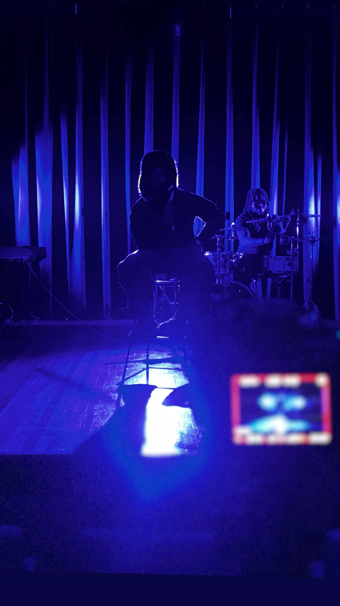 Silhouette of a person playing guitar on stage with blue lighting and curtains, audience in foreground, camera recording.