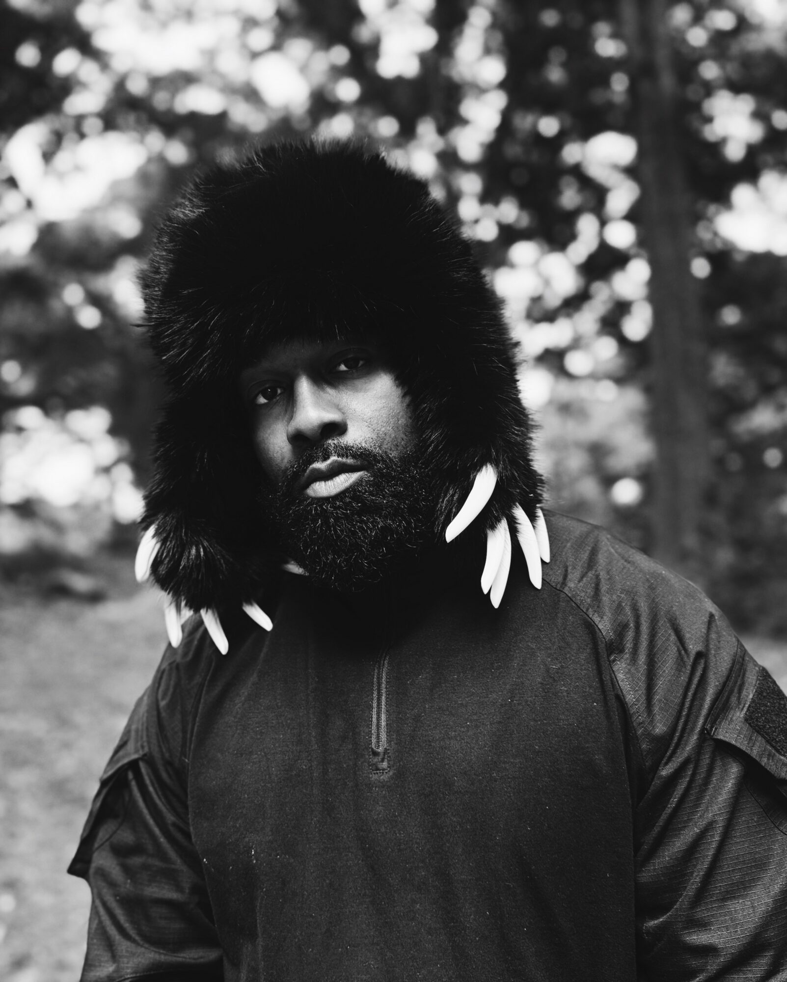 Man with a large fur hat and feather earrings outdoors, looking at the camera, black and white photo.
