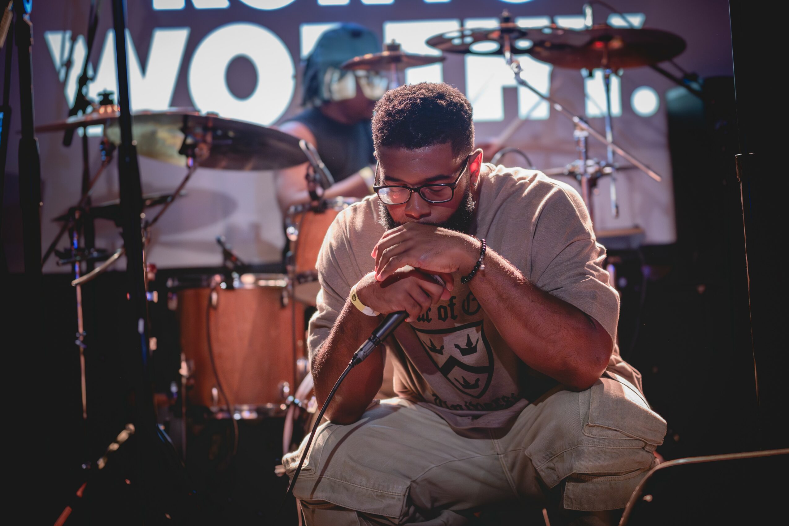 A man with glasses and a beige shirt sits on stage with a microphone, with a drummer and drum set in the background.