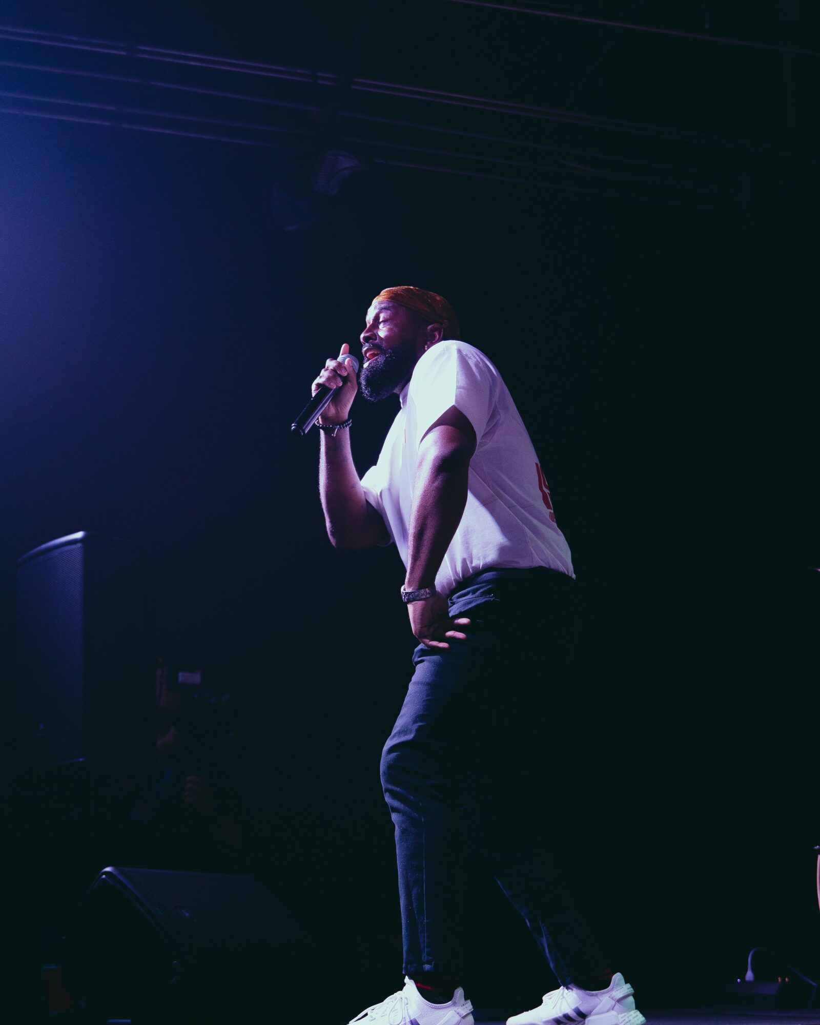 Person singing into a microphone on stage with dark background, wearing a white shirt and dark pants.