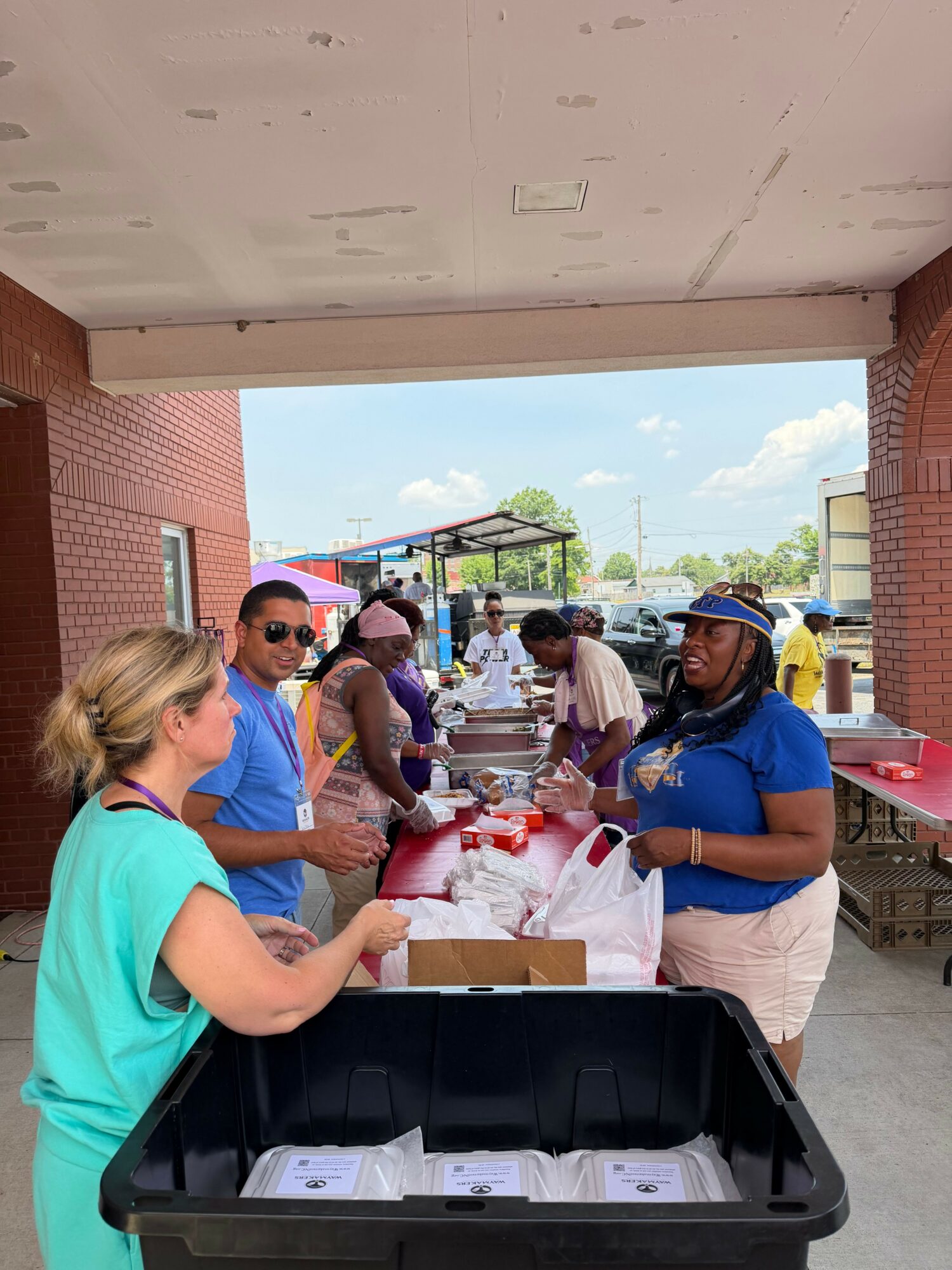 People gather around a table outdoors, with a woman in a green shirt and a woman in a blue shirt, under a brick structure.