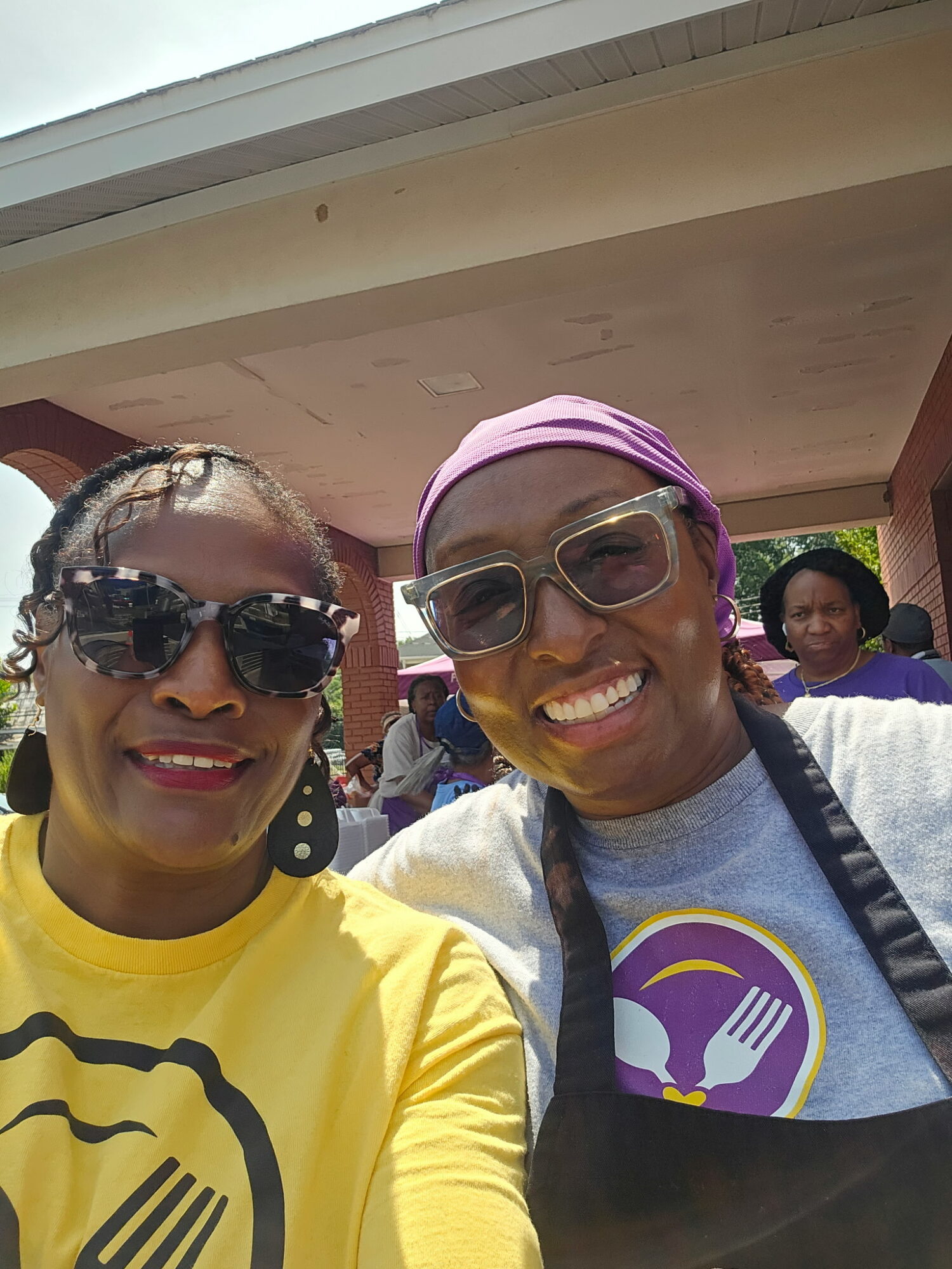 Two women smiling outdoors, one wearing sunglasses and a yellow shirt, the other wearing glasses, a purple headscarf, and a gray shirt with a logo.