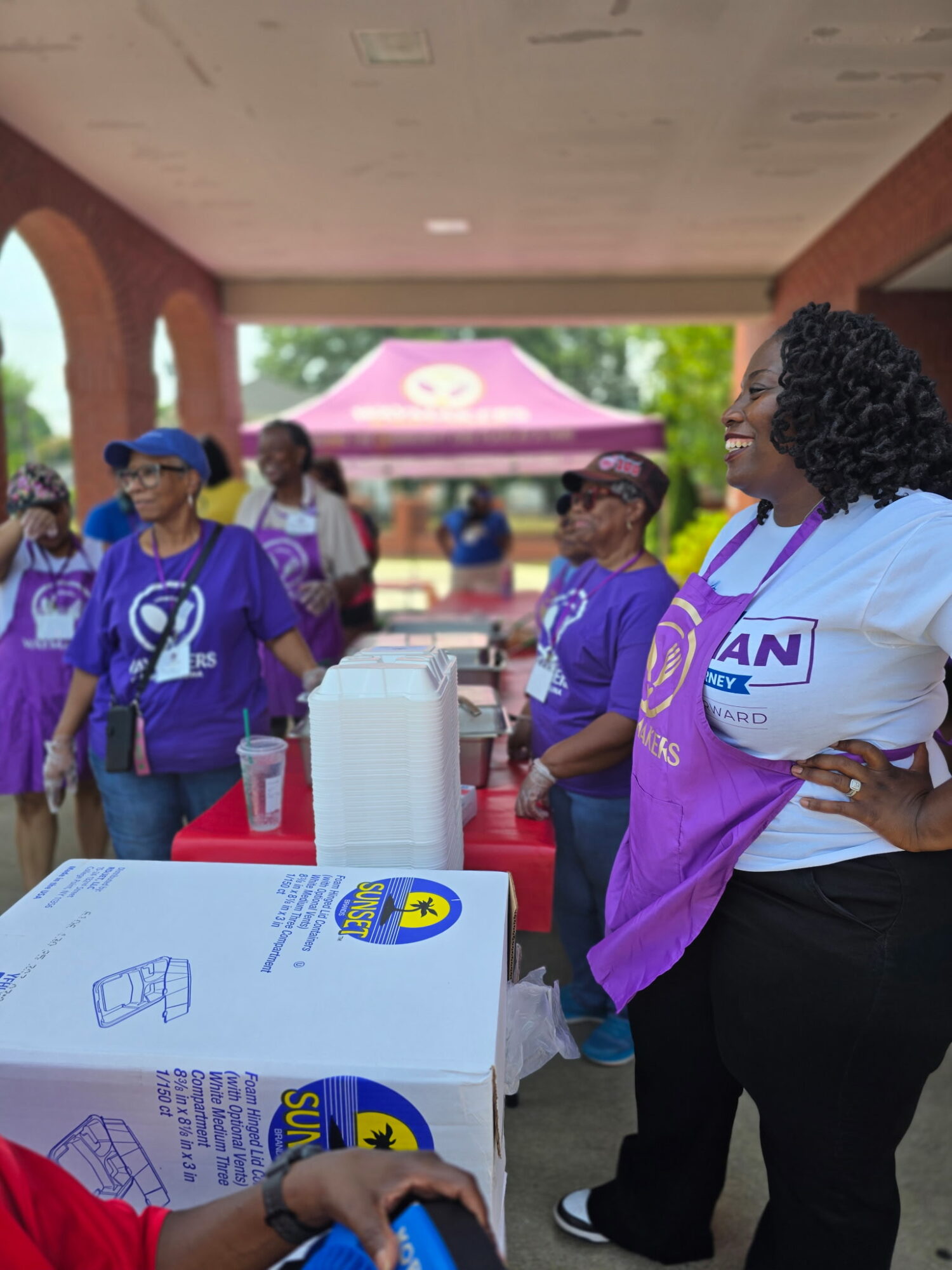 Group of people wearing purple shirts gathered outdoors near a table with a white box, under a pink canopy, with trees in background.
