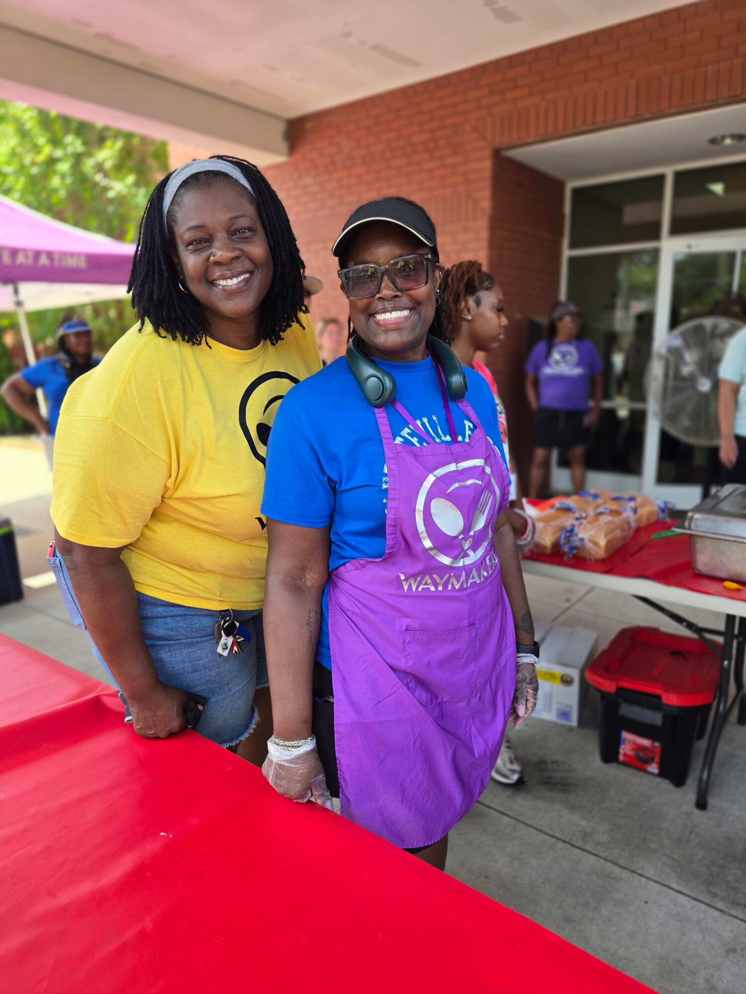 Two smiling women standing outdoors near a red table, one wearing a yellow shirt and the other in a blue shirt with a purple apron, with people and a building in the background.