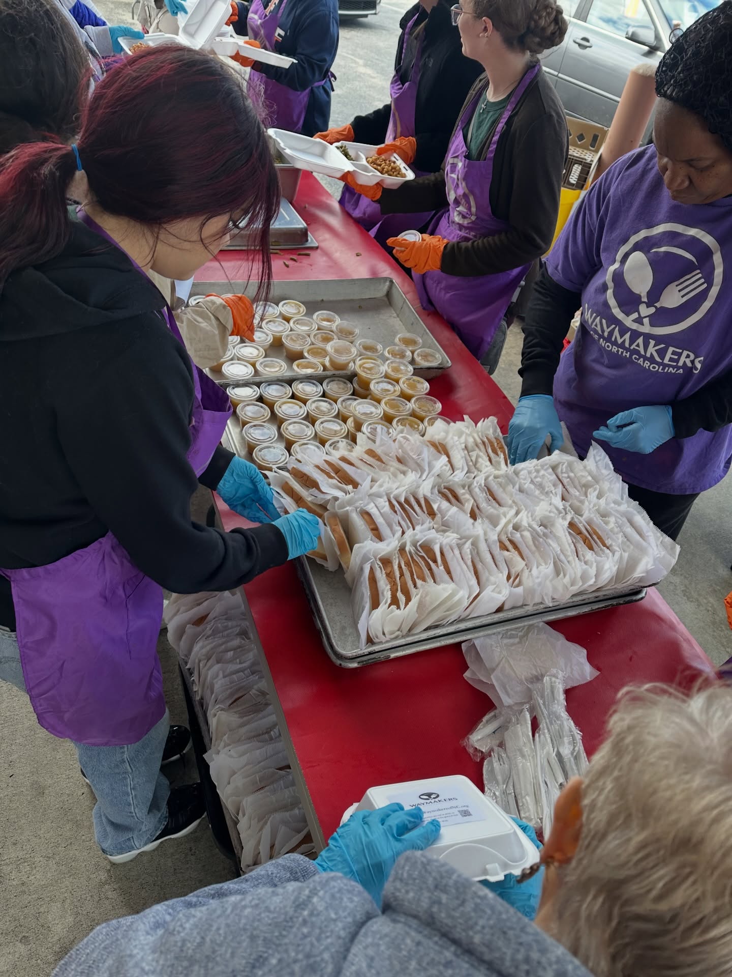 People preparing and packaging food items at a table outdoors, wearing purple aprons and gloves.