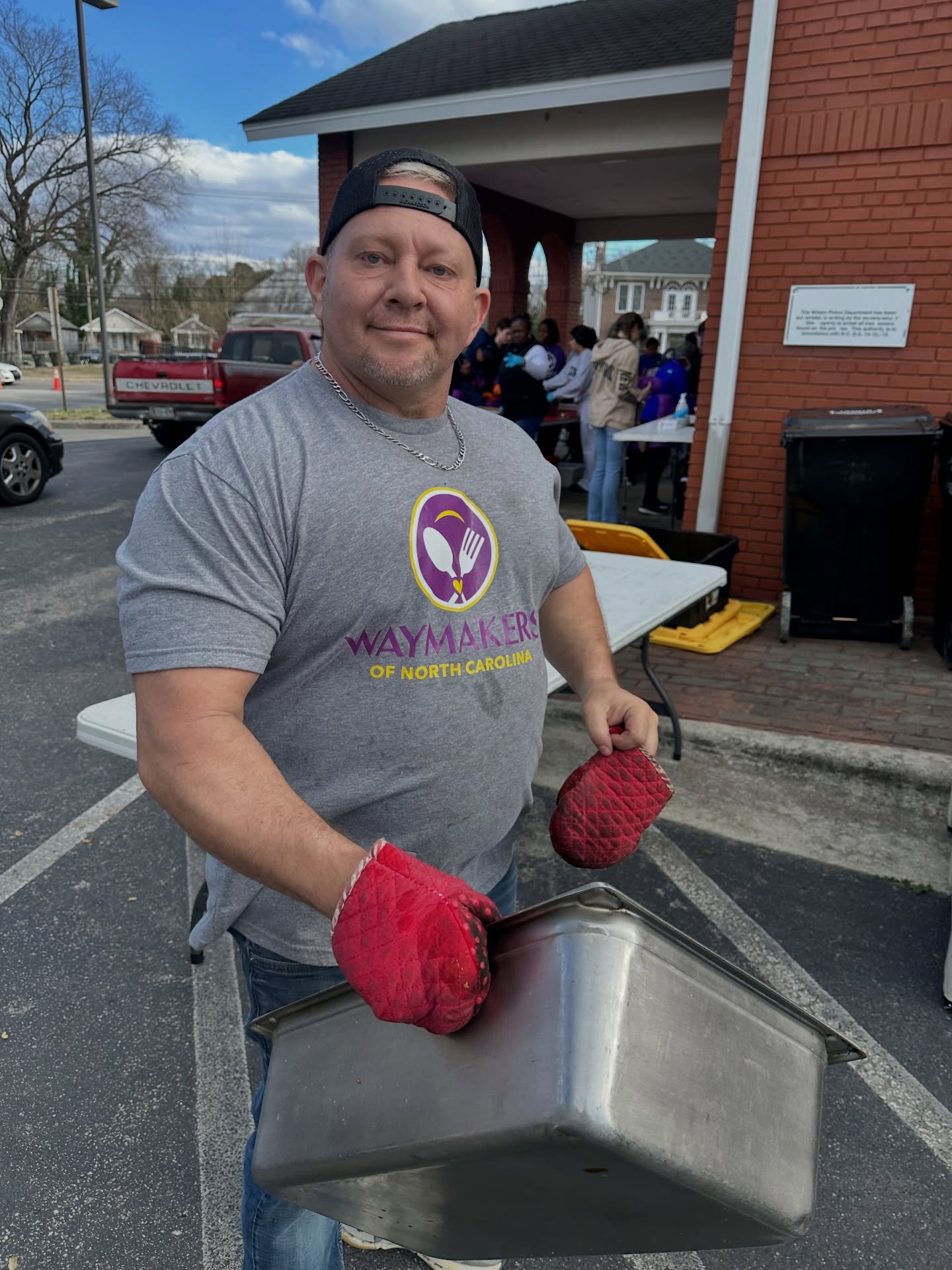 Man wearing a gray T-shirt and red gloves holding a metal container outside a building with people in the background.