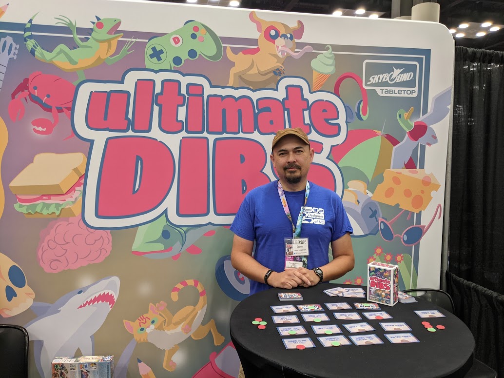 Man standing behind a table with cards at a booth for Ultimate Dibs, colorful cartoon characters in background.