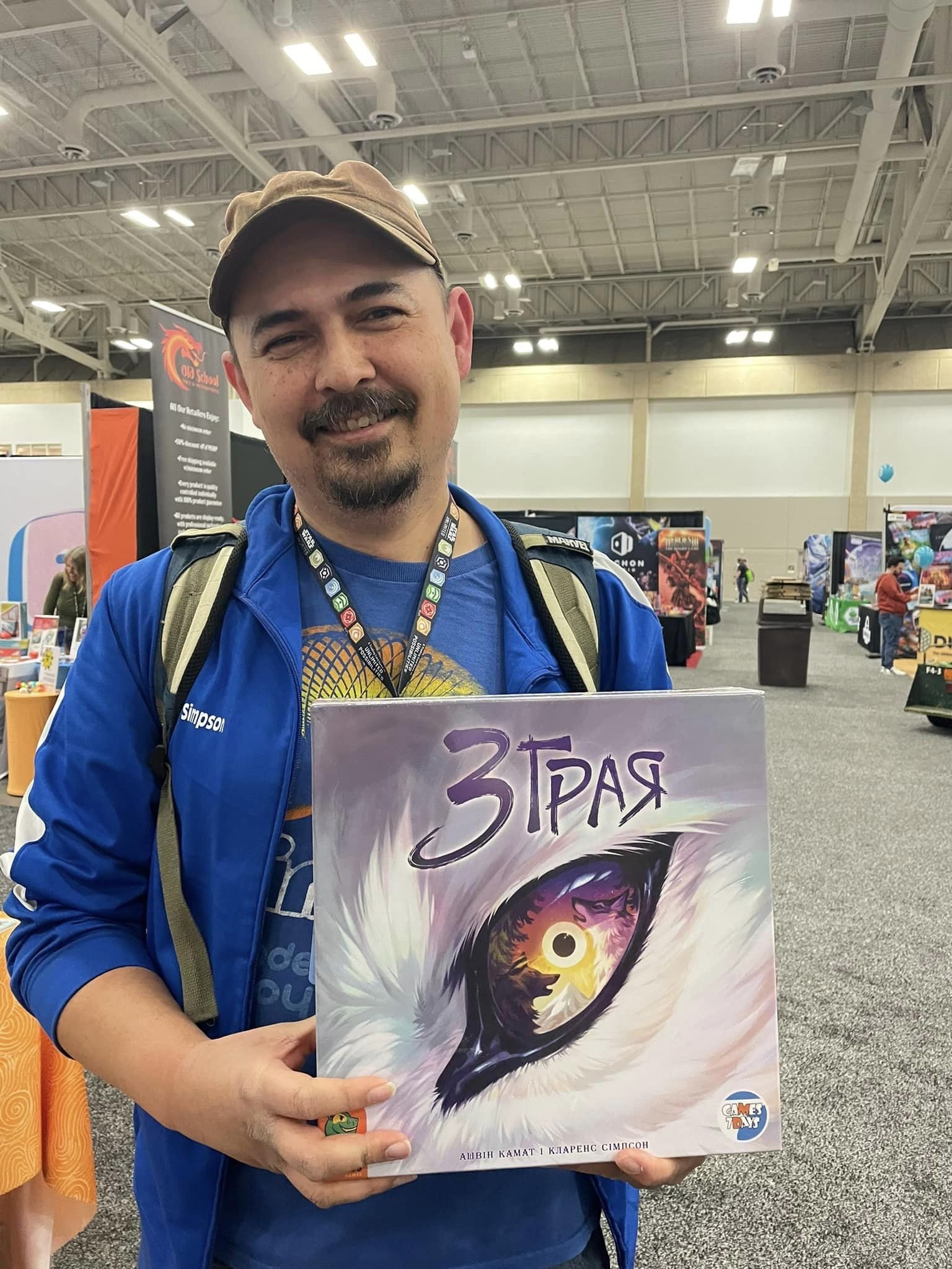 Man with a beard and cap holding a colorful art piece at an indoor event, with booths and displays in the background.