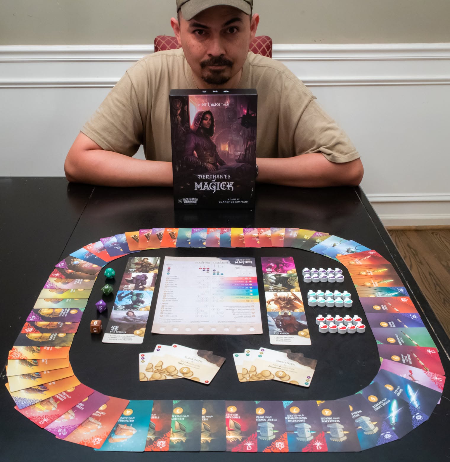 Person sitting at a table with a board game, game components, and a game box, in a room with white wall paneling.