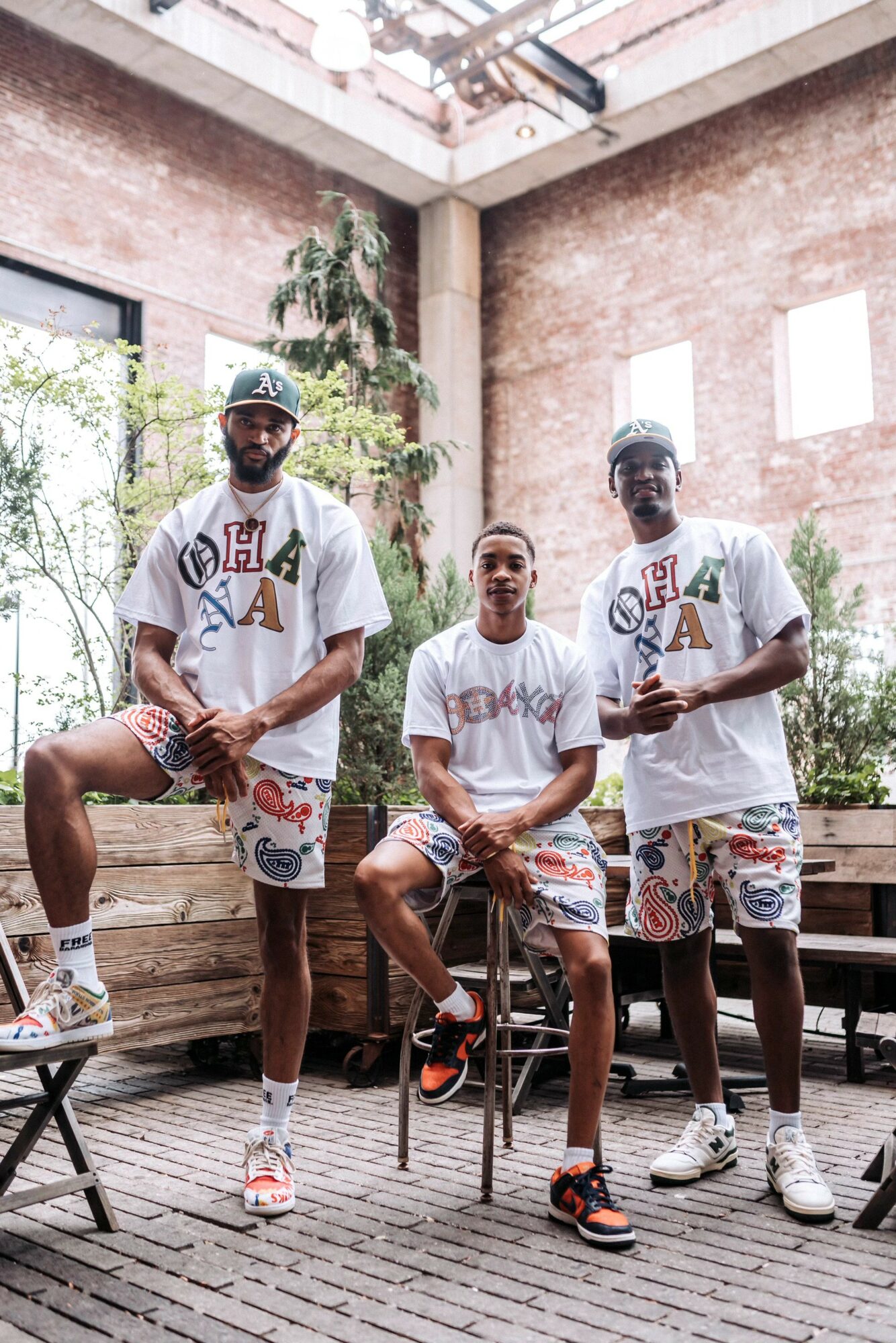 Three men in casual clothing and hats pose indoors with brick walls and plants in the background.