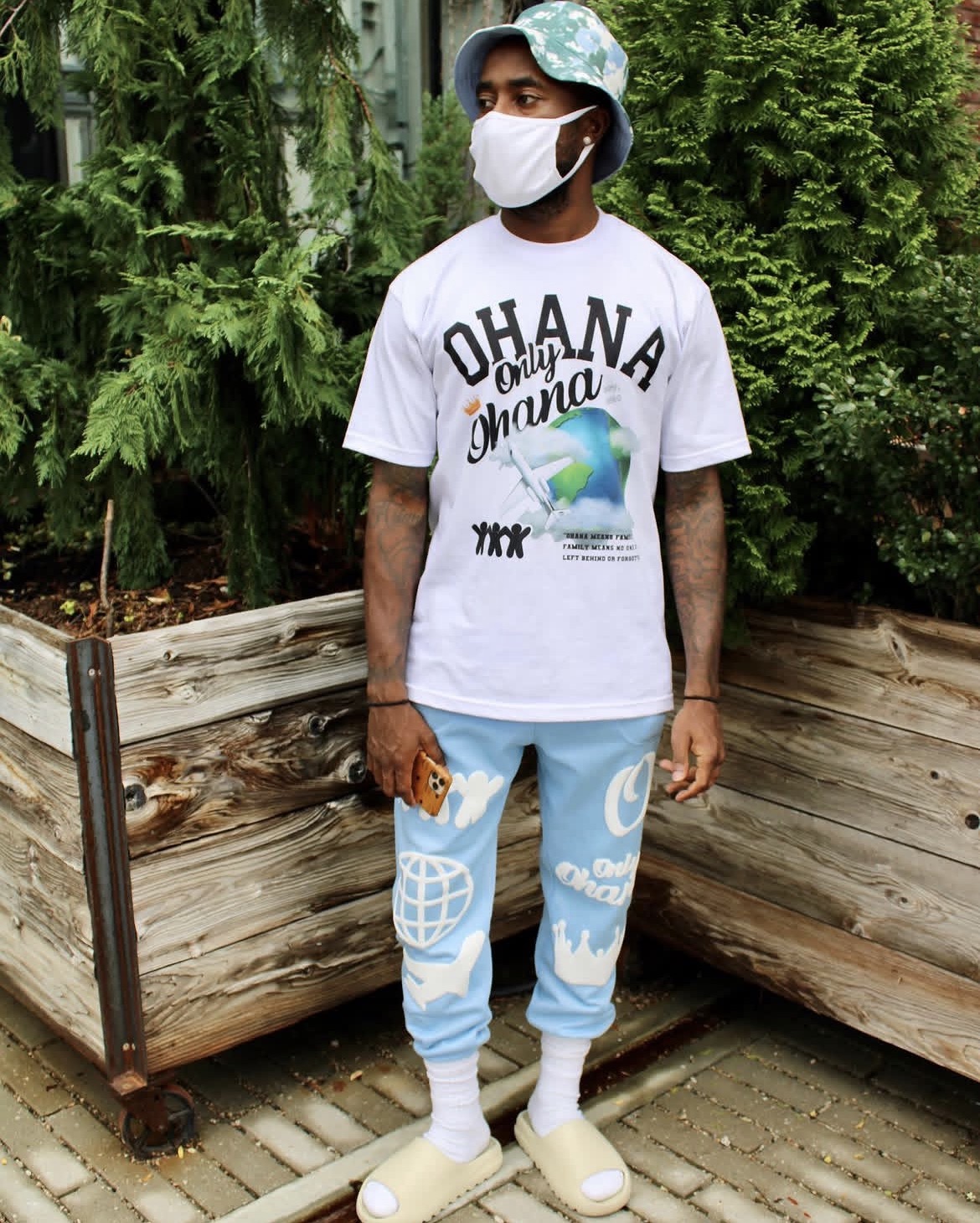 Person wearing a white mask, bucket hat, white T-shirt with text and graphic, light blue pants with white patterns, standing outdoors