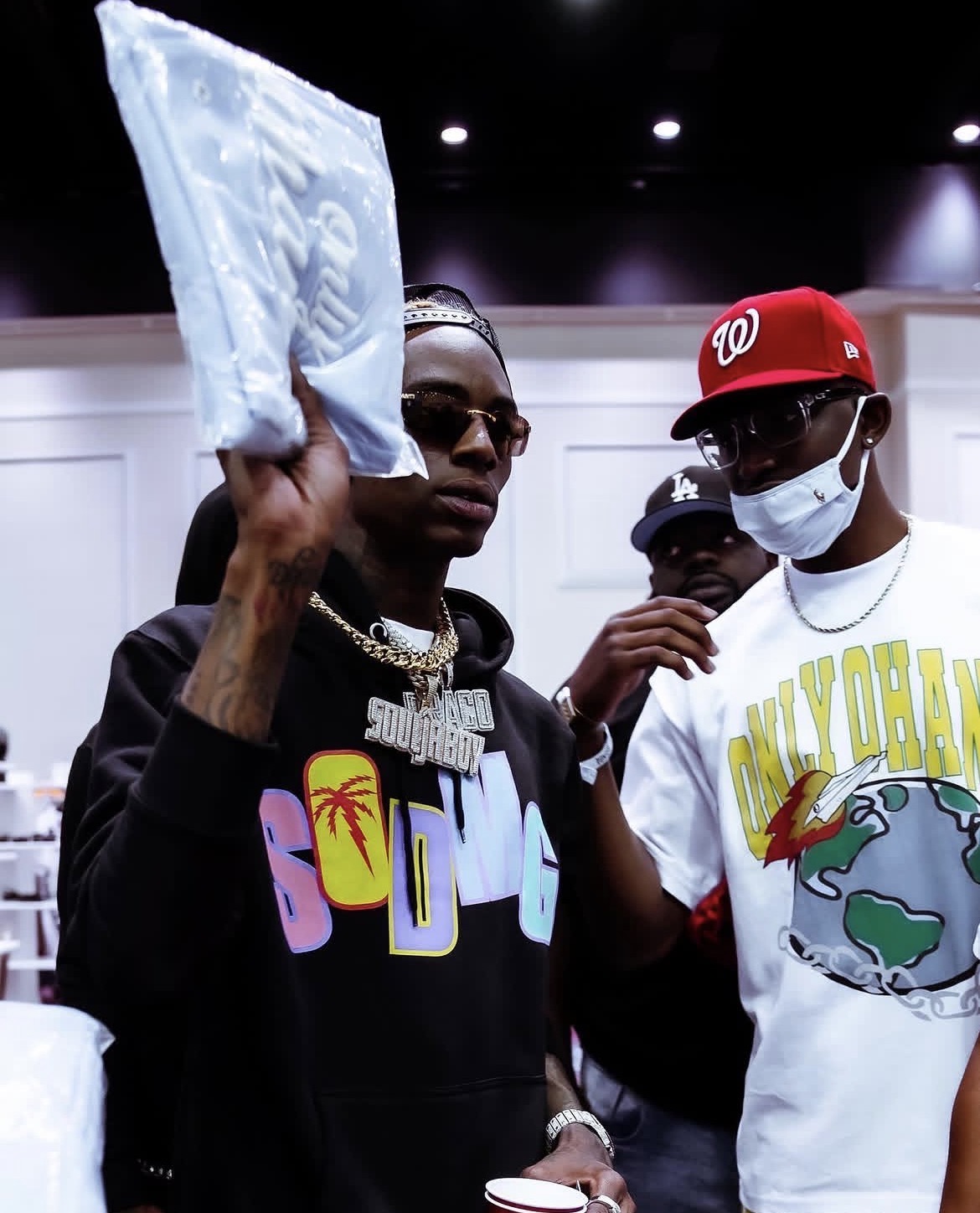 Person holding a large white object, wearing sunglasses, a black hoodie with colorful text, and jewelry, in a group setting.